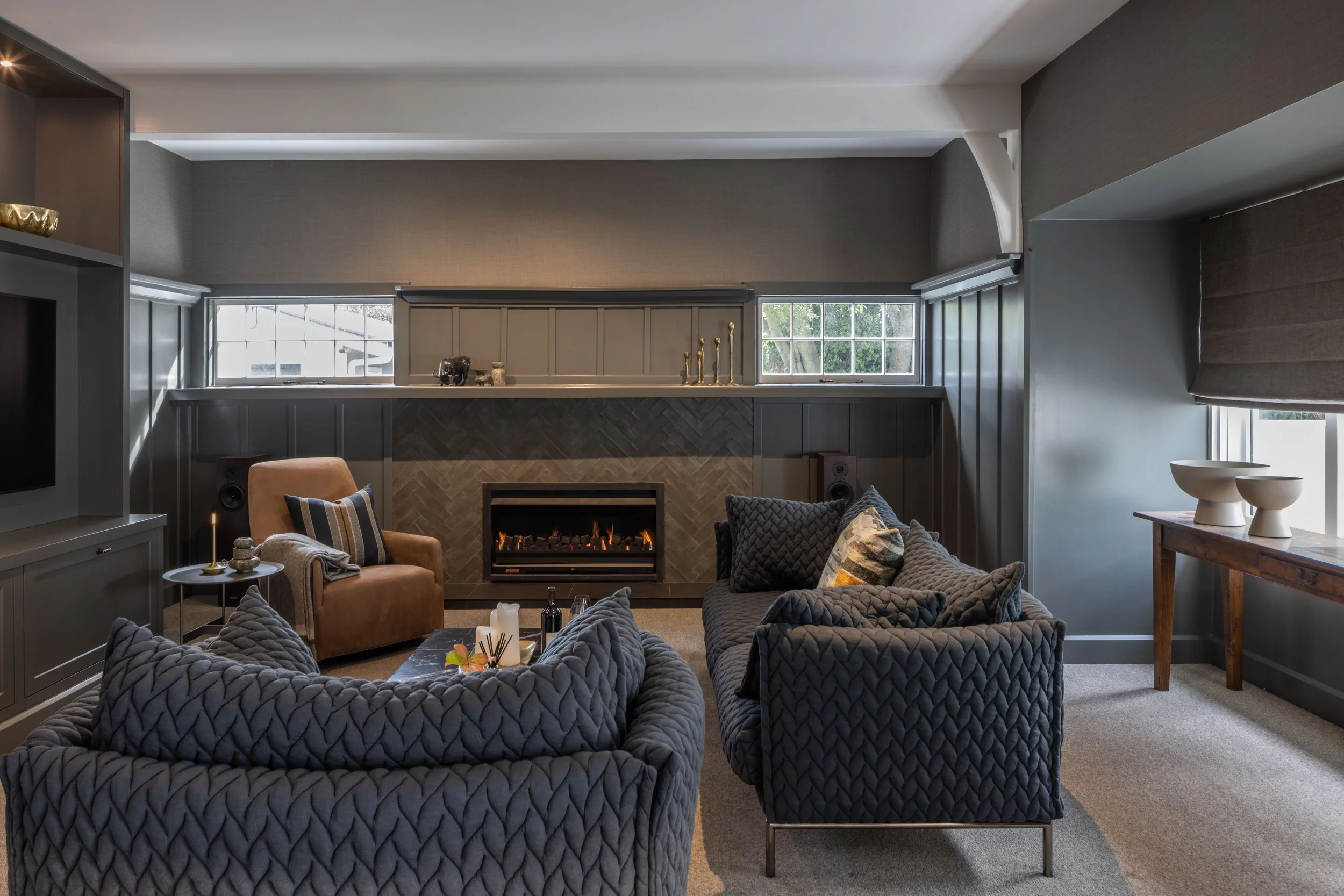 A cozy living room with a fireplace, gray upholstered armchair and sofa, window with gray blinds, and a wooden console table with decorative items.