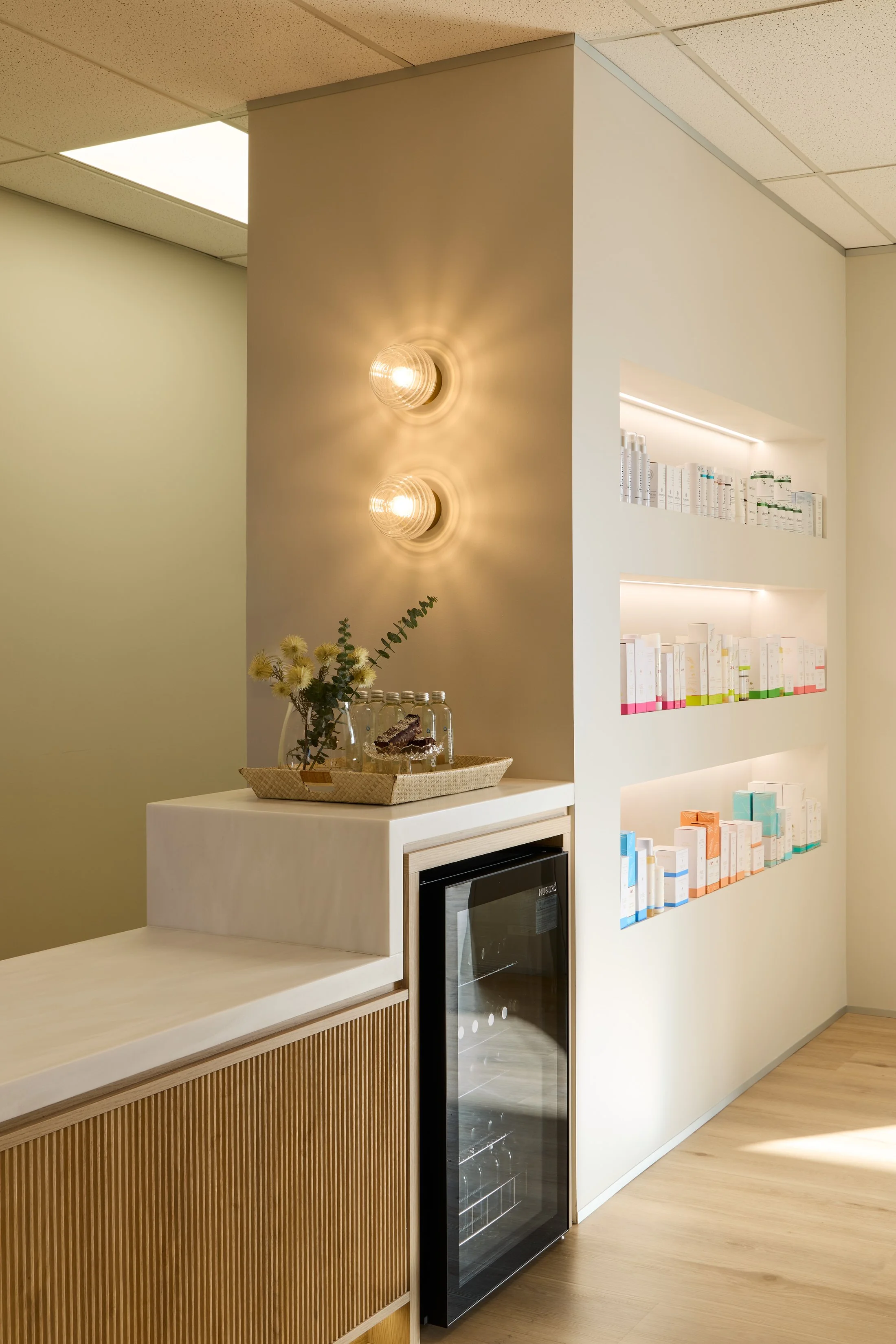 Interior view of a modern room with a corner featuring two warm wall lights, a white wall with built-in shelves holding skincare or cosmetic products, a small countertop with a flower vase and snacks, a mini fridge, and a microwave above a wooden cabinet.