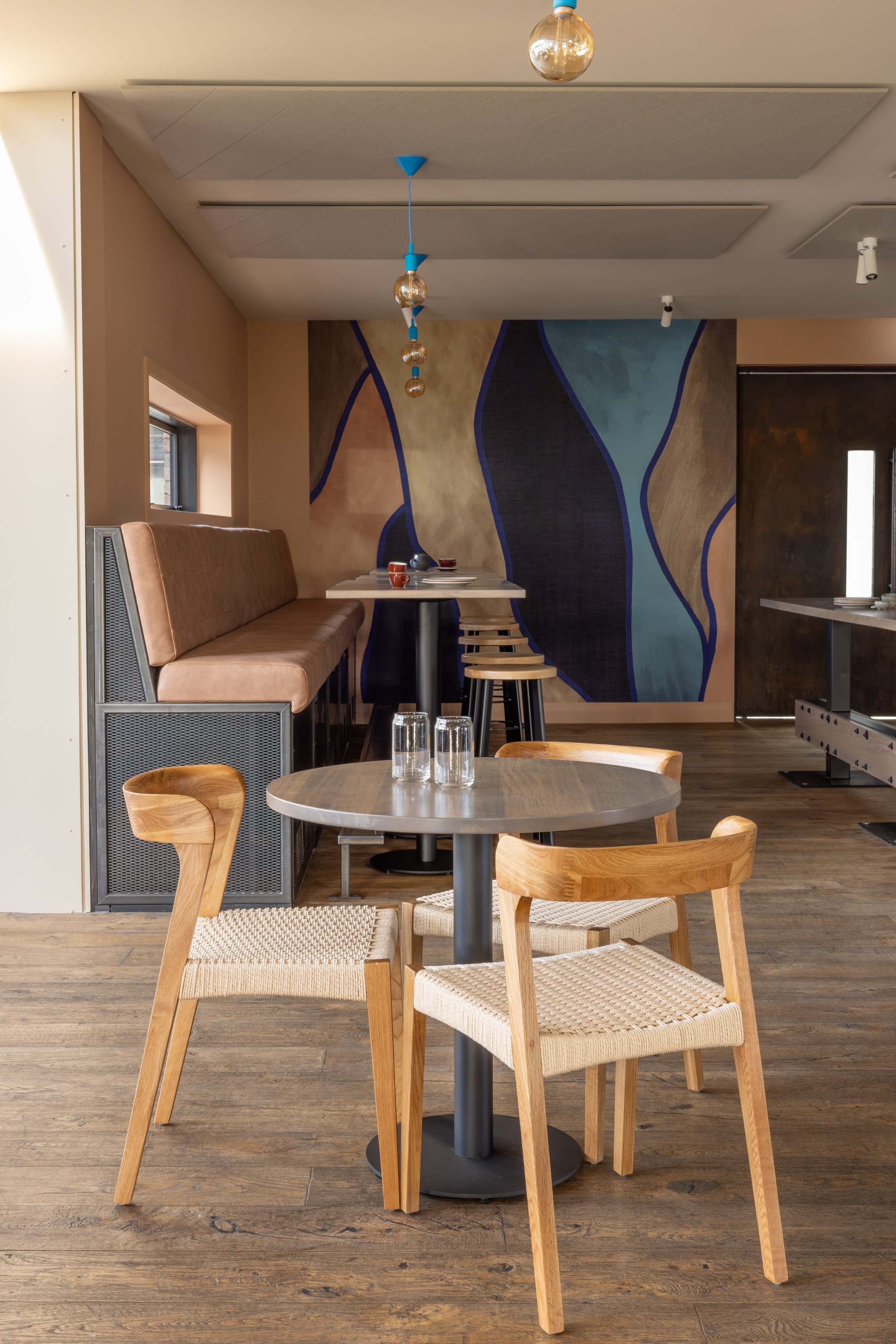 Interior of a modern restaurant with wooden floors, wooden chairs with woven seats, a table set with glasses, and a wall with colorful abstract art. There are hanging light fixtures and a long padded bench along the wall.