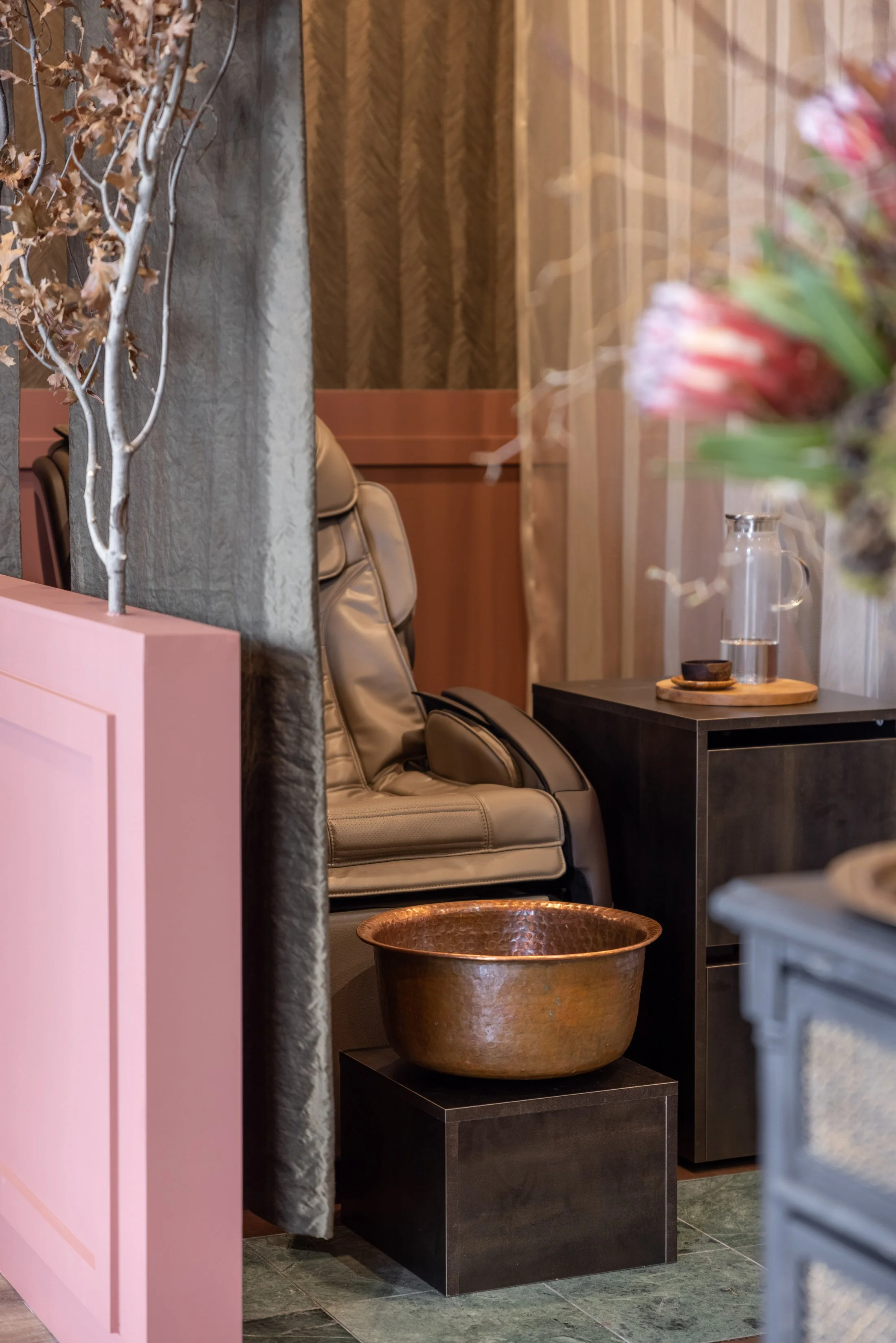 A cozy indoor space with beige leather armchairs, a black side table with a wooden tray, a glass pitcher, and a cup. A copper basin is on the floor, and a pink partition with decorative dried plants separates the seating area from the foreground. The wall behind has textured wood paneling.