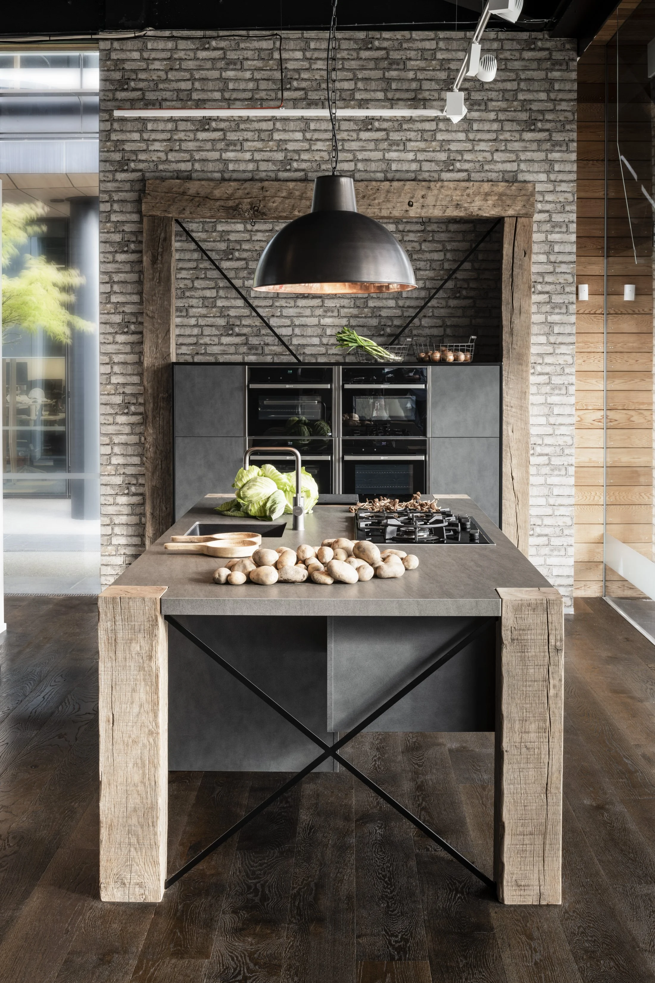 A modern kitchen with a large island topped with potatoes, lettuce, and mushrooms, a black pendant light above, and a brick wall backdrop with wooden accents.