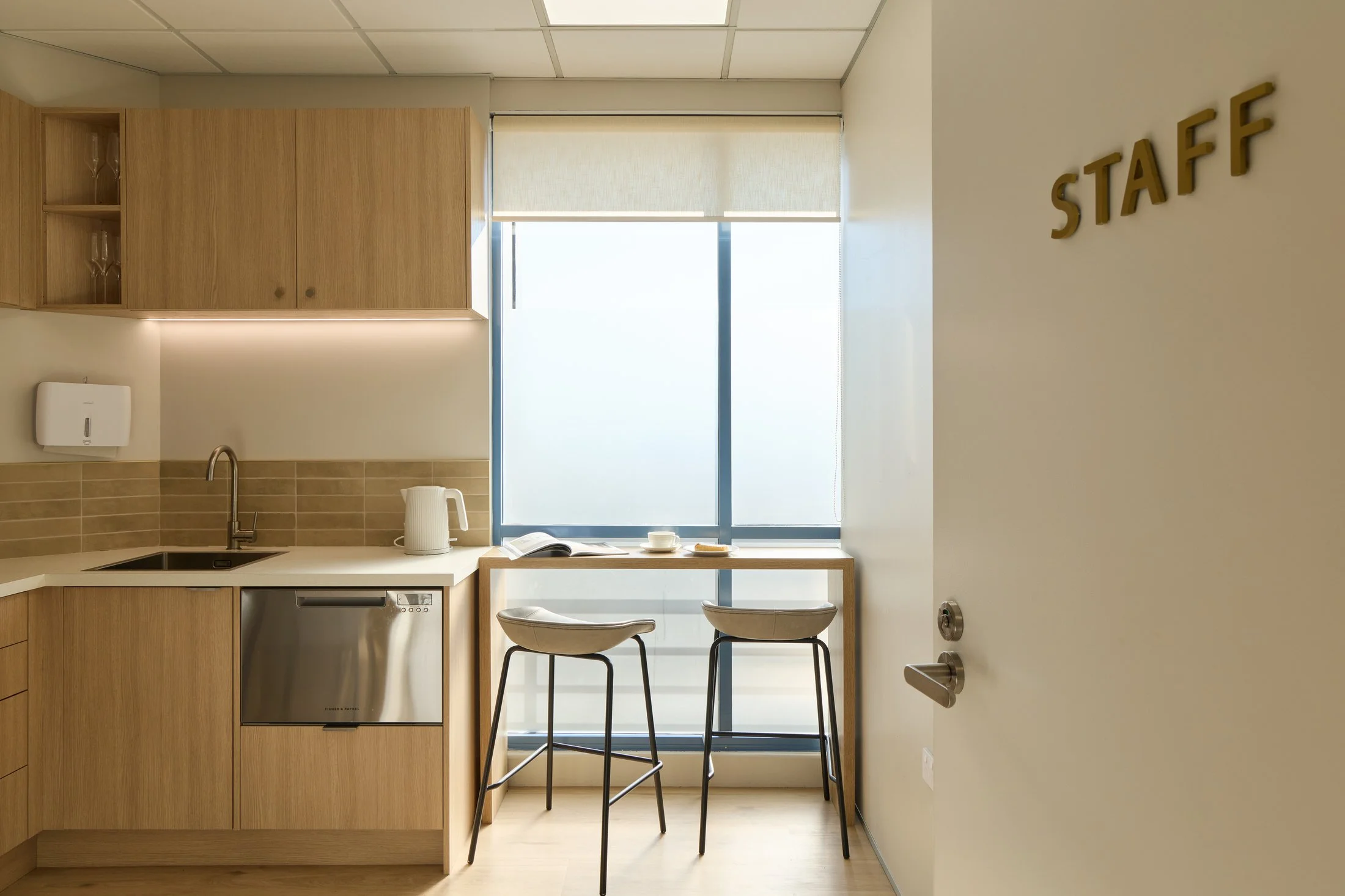 Interior of a modern break room or kitchenette with wooden cabinets, a window with blinds, a small table holding a book, cup, and saucer, and two black metal chairs. A wall sign says 'STAFF'.