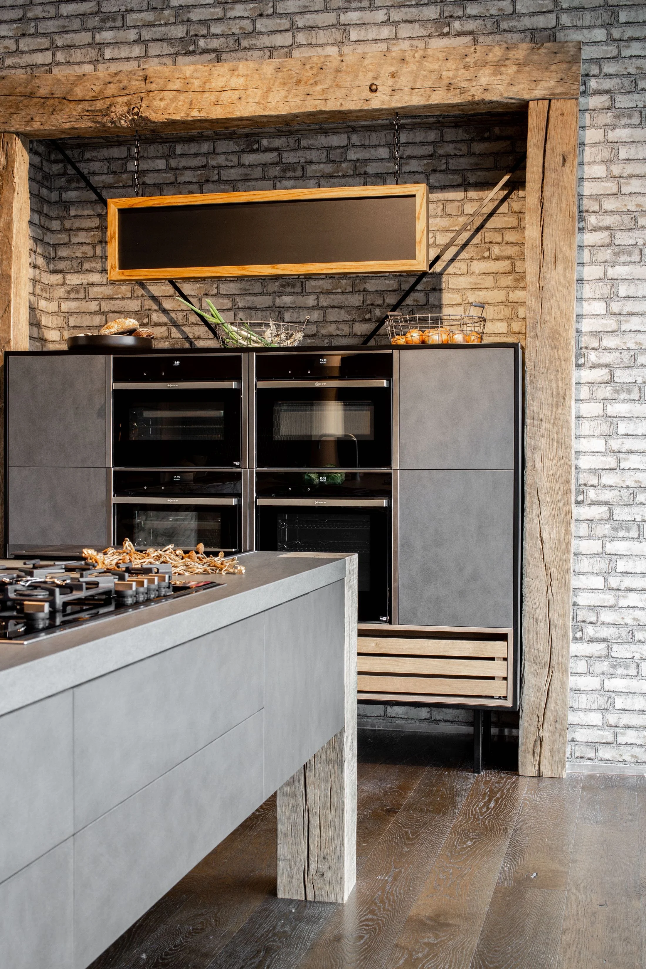 Modern kitchen with gray cabinetry, stainless steel ovens, a stone gray island countertop with dried herbs and miscellaneous items, and a brick wall with wooden accents and a suspended chalkboard or sign. There are onions in a wire basket and a black pan on the counter, with wooden flooring.