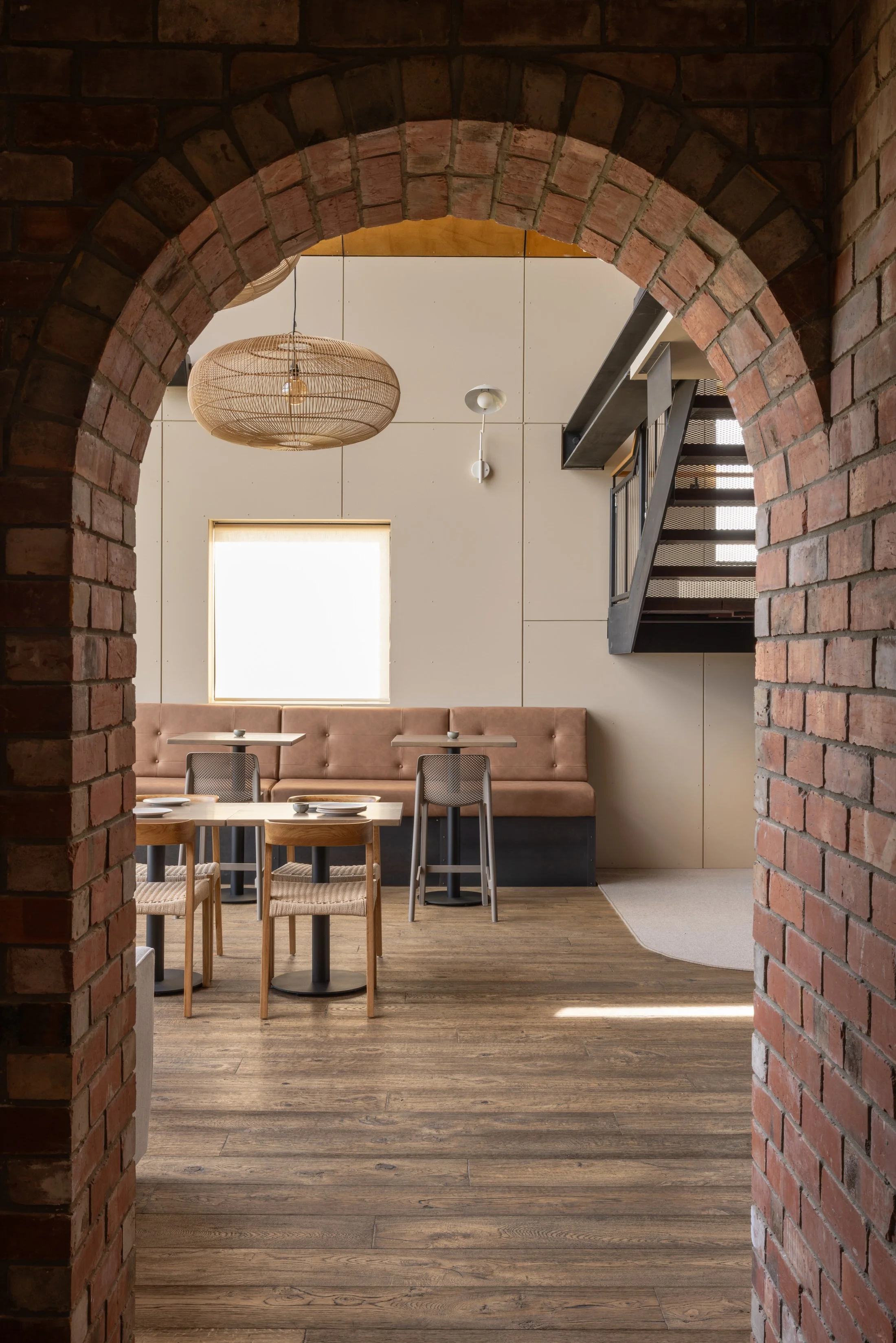 Interior of a modern restaurant or cafe seen through a brick archway, with wooden floors, a beige cushioned bench, tables with chairs, a large window, and a metal staircase.