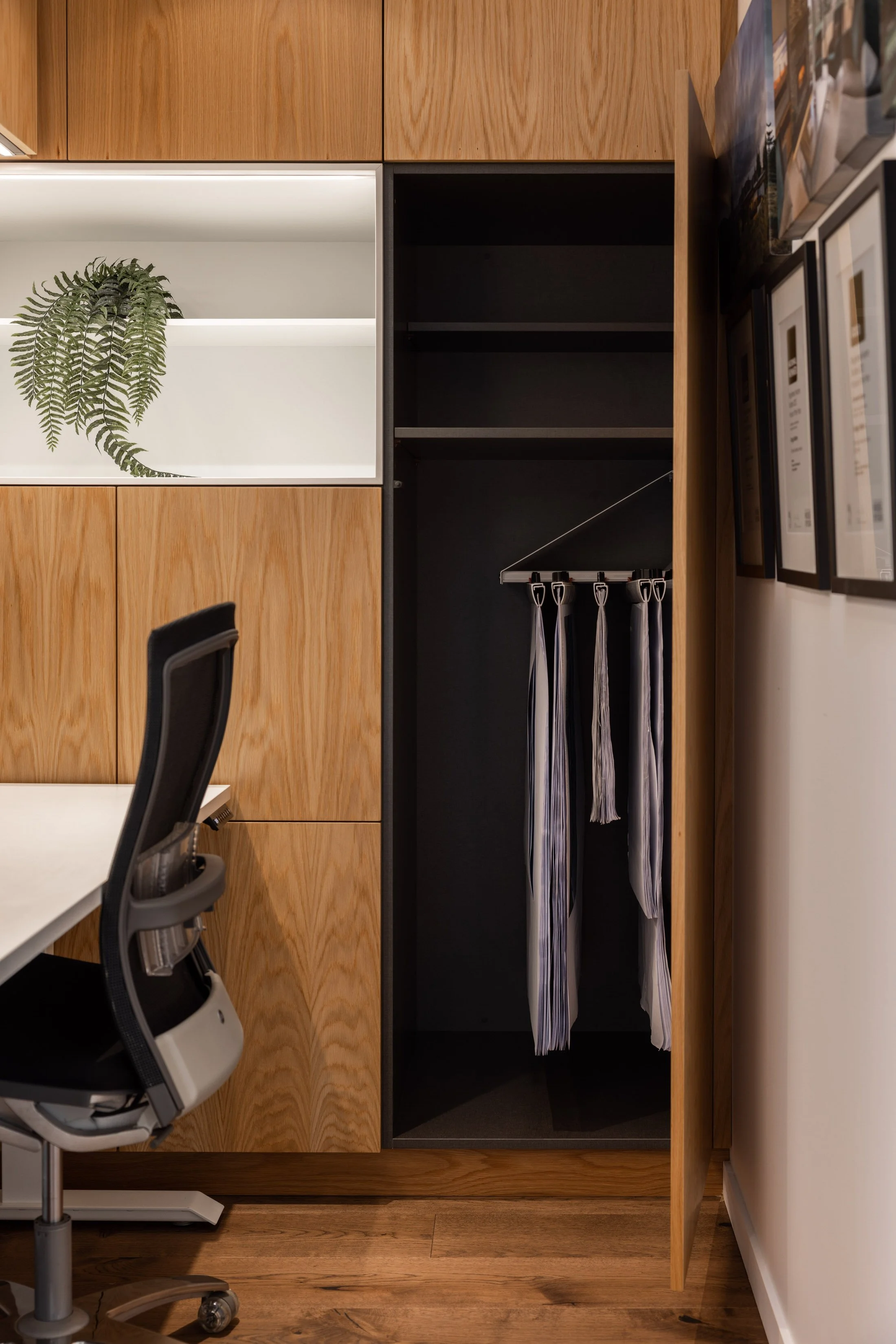 Interior office space with wooden wall cabinets, open black storage closet with hanging ties, a desk with a white surface, and an office chair with a mesh back and helmet on top. Framed pictures hang on the light-colored wall.