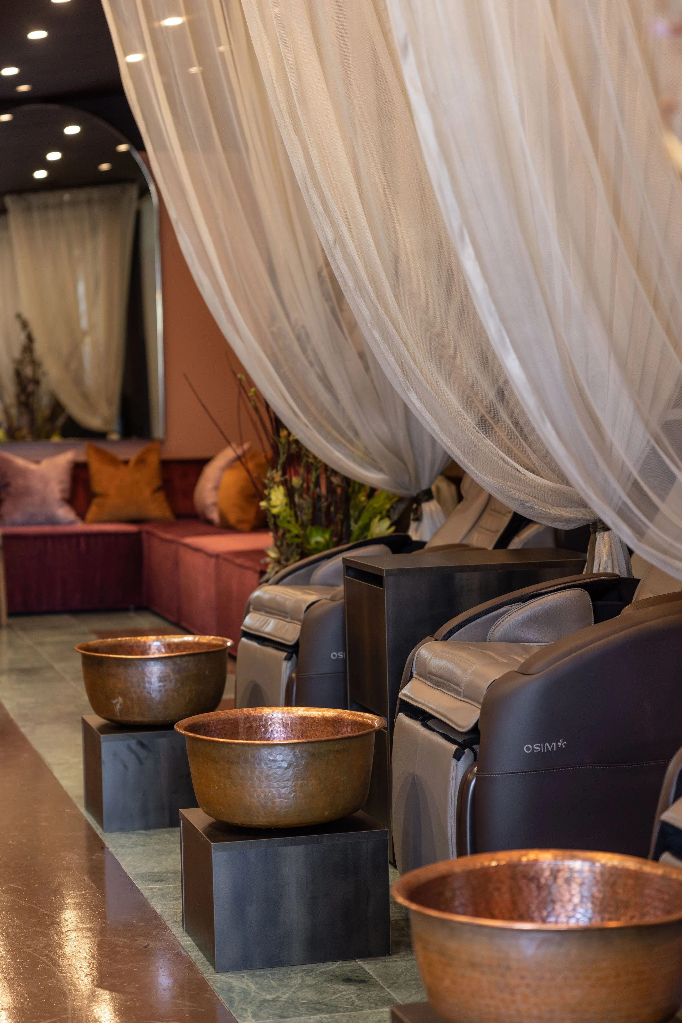 A row of massage chairs with large copper bowls in front, positioned against a wall with beige curtains, in a spa or wellness center setting.