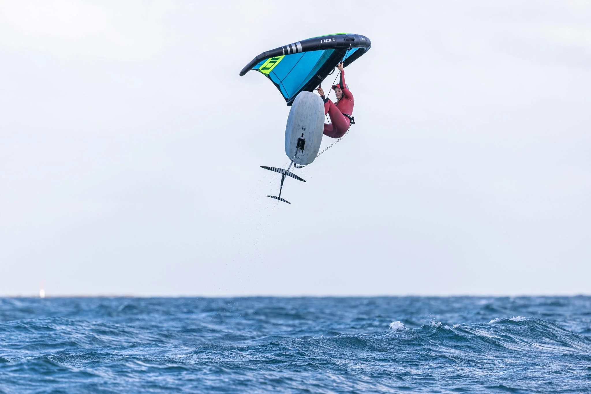 Person in pink wetsuit performing a high jump on water with a wing sail windsurfing rig.