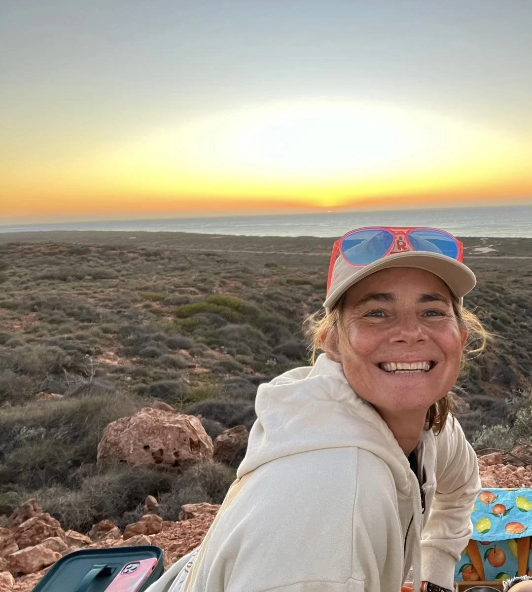 A woman smiling outdoors during sunset by the ocean, wearing a light-colored hoodie, a beige cap, and red sunglasses with blue lenses, with rocky terrain and sparse vegetation in the background.