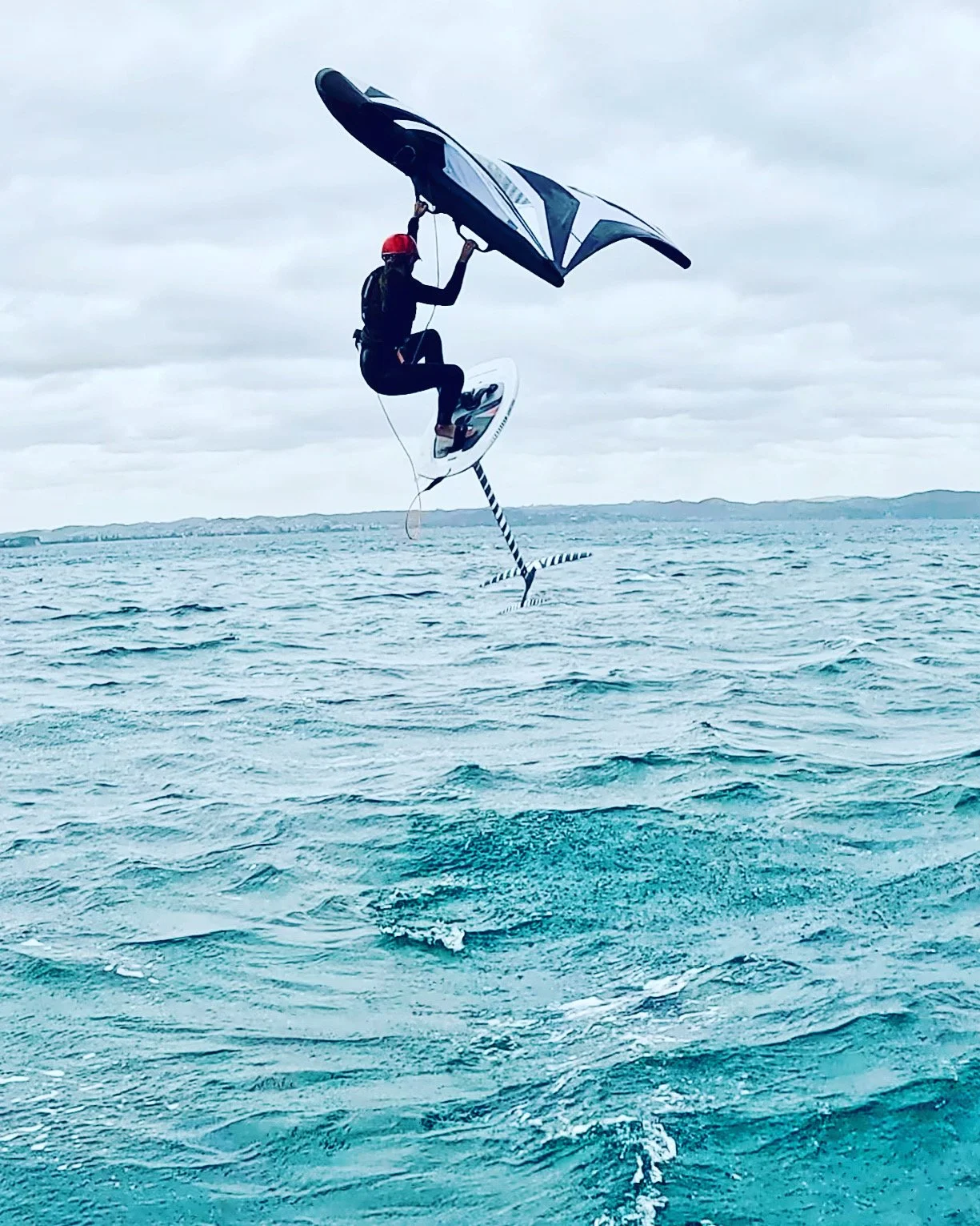 Person wearing a red helmet and black wetsuit, holding a wing sail, riding a foil board on the ocean during overcast weather.