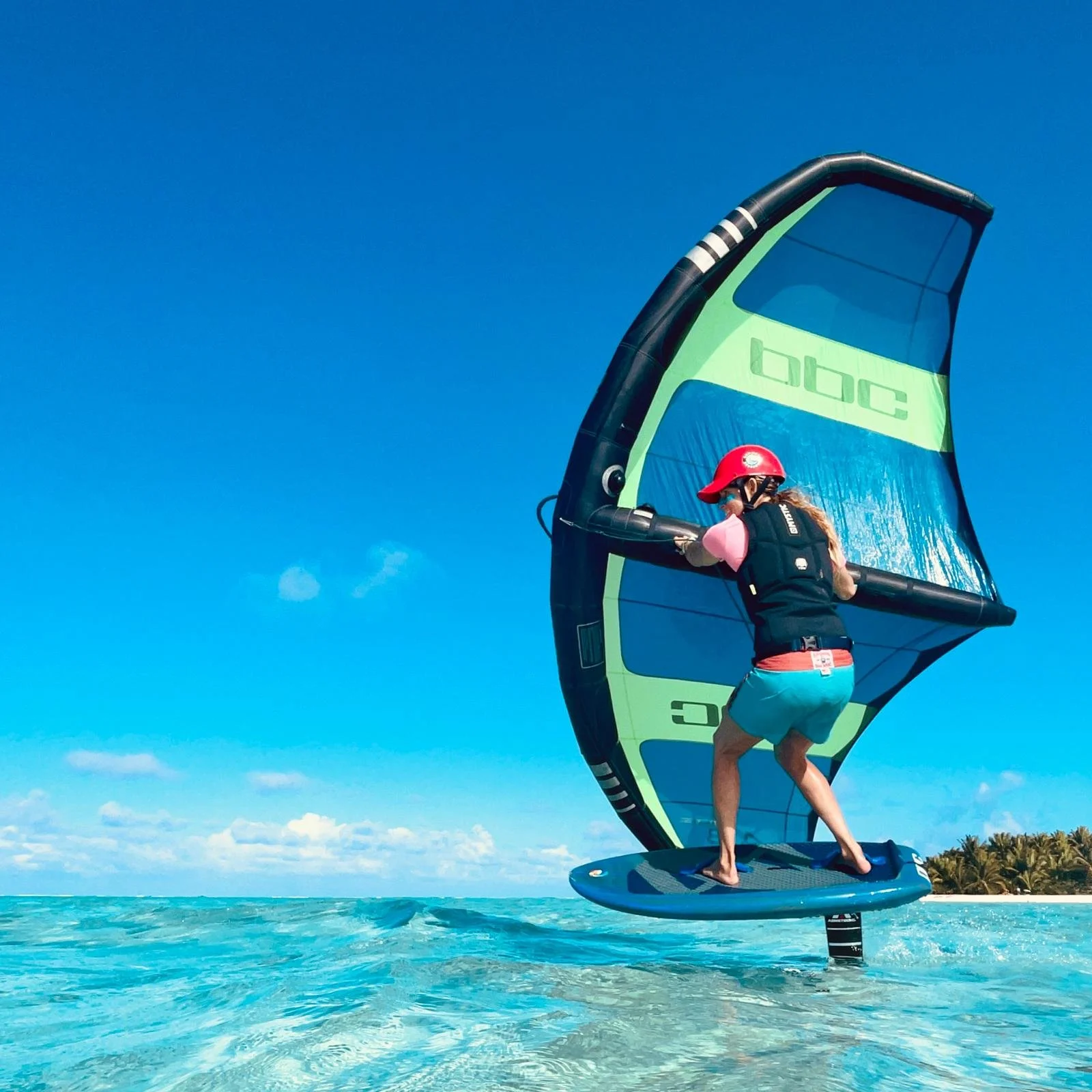 A person riding a hydrofoil wing surfboard with a wing sail on clear blue water with a small island and palm trees in the distance.