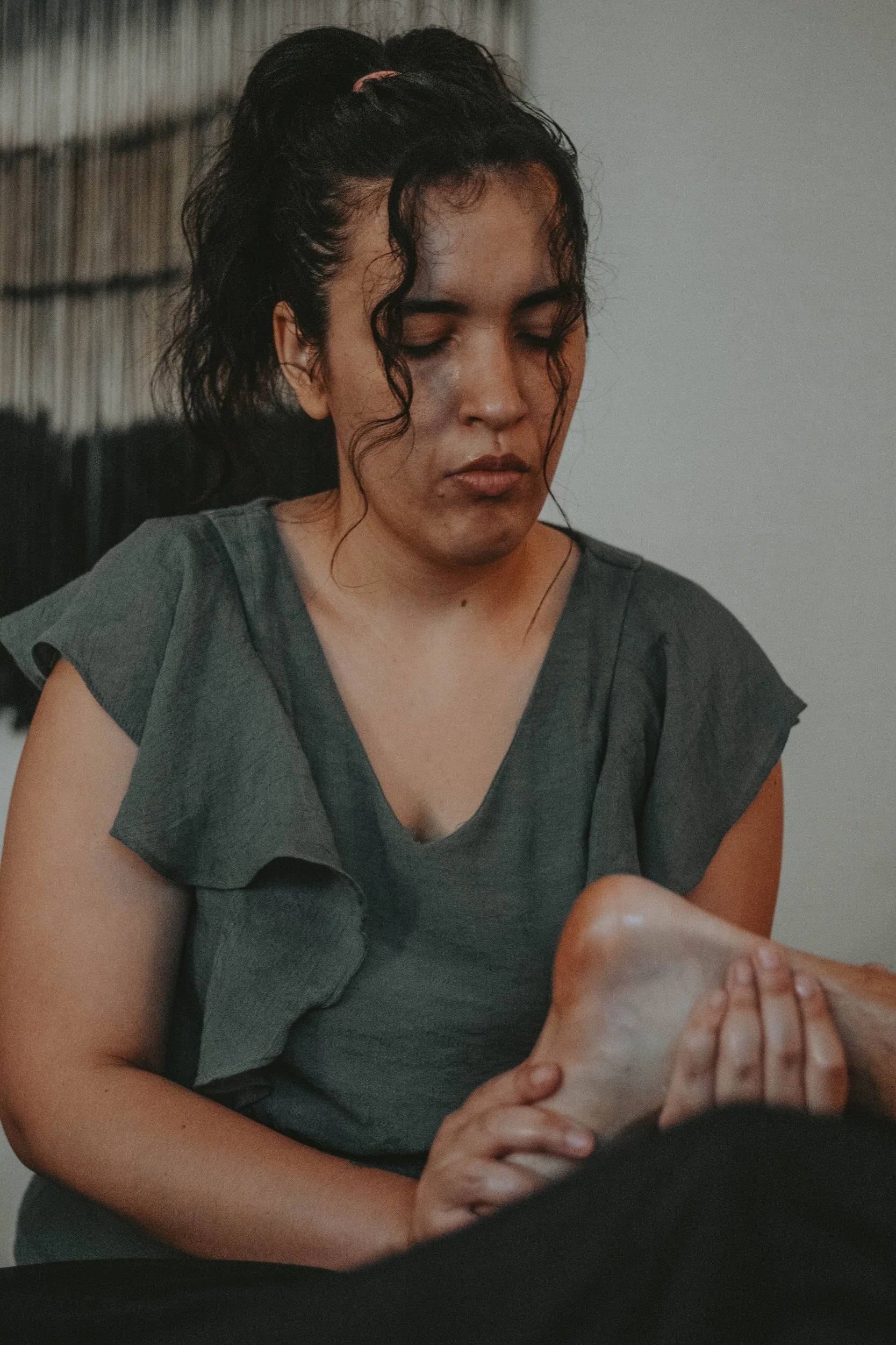 A woman with dark, curly hair tied back, wearing a gray top, is sitting and looking downward, with her eyes closed or looking down, possibly focusing or resting.