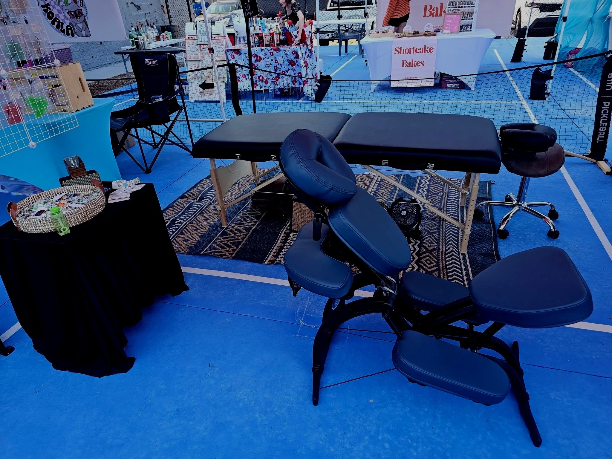 An outdoor event setup on a blue sports court with vendor tables, including a massage table with a black cushion, a table with baked goods labeled 'Shortcake Bakes,' and other booths selling various items. There is some equipment and chairs around, and a net barrier separates the vendor area from the courts.
