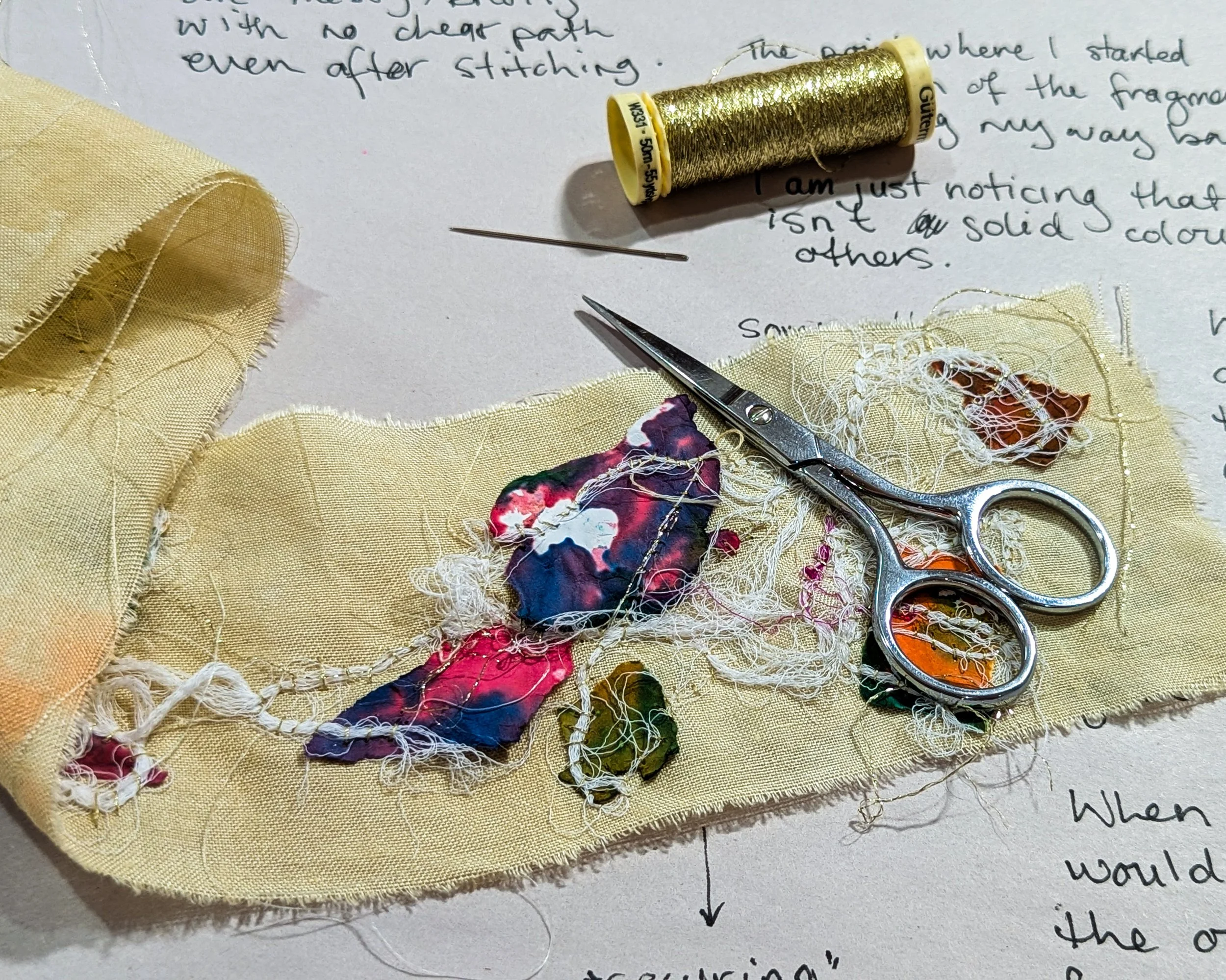 Fabric with stitched fragments, scissors, and a spool of gold thread resting on handwritten notes.