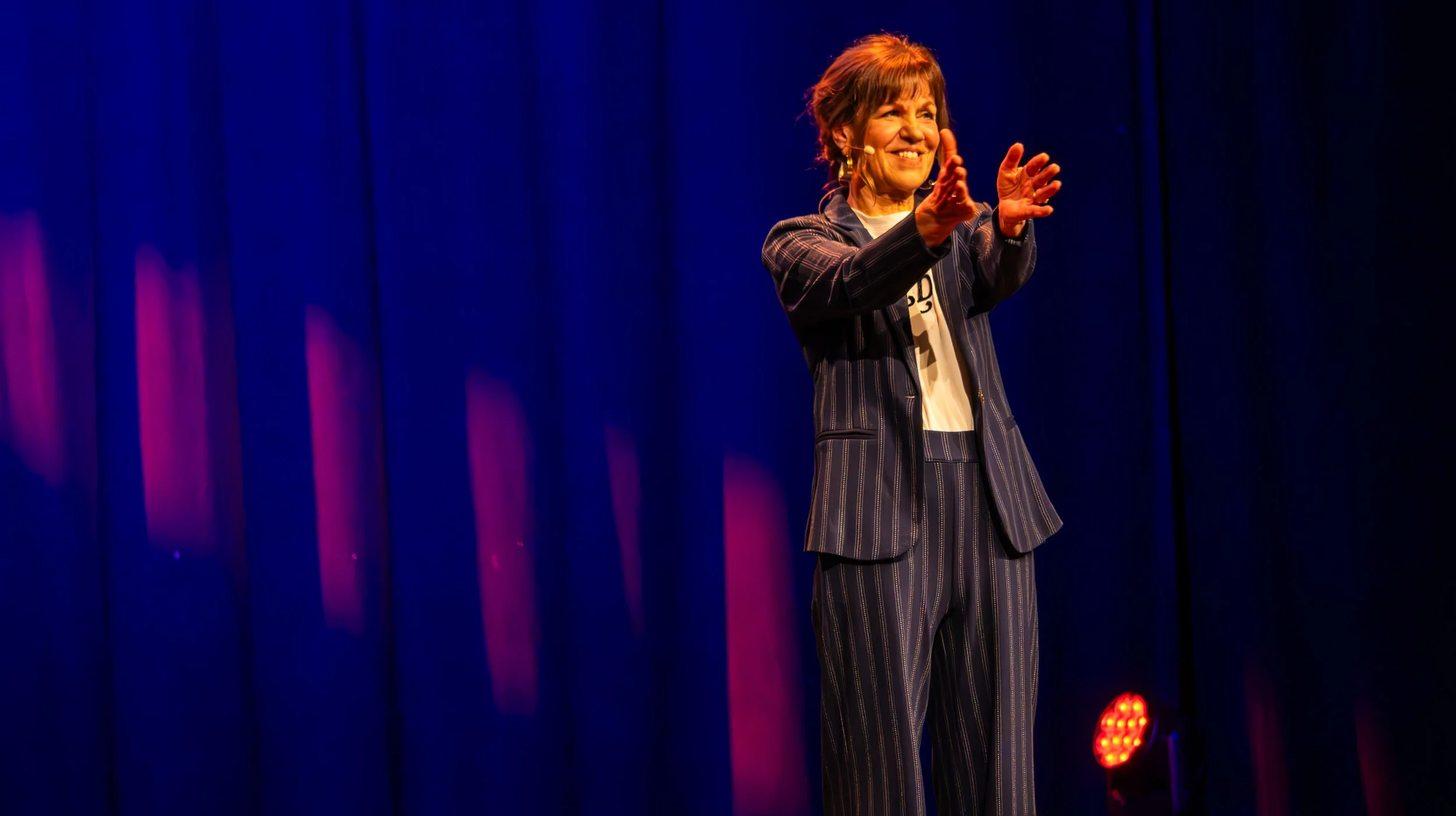 Een glimlachende vrouw op een podium met een donkerblauwe achtergrond en rood theaterlicht, gekleed in een gestripeerd pak.