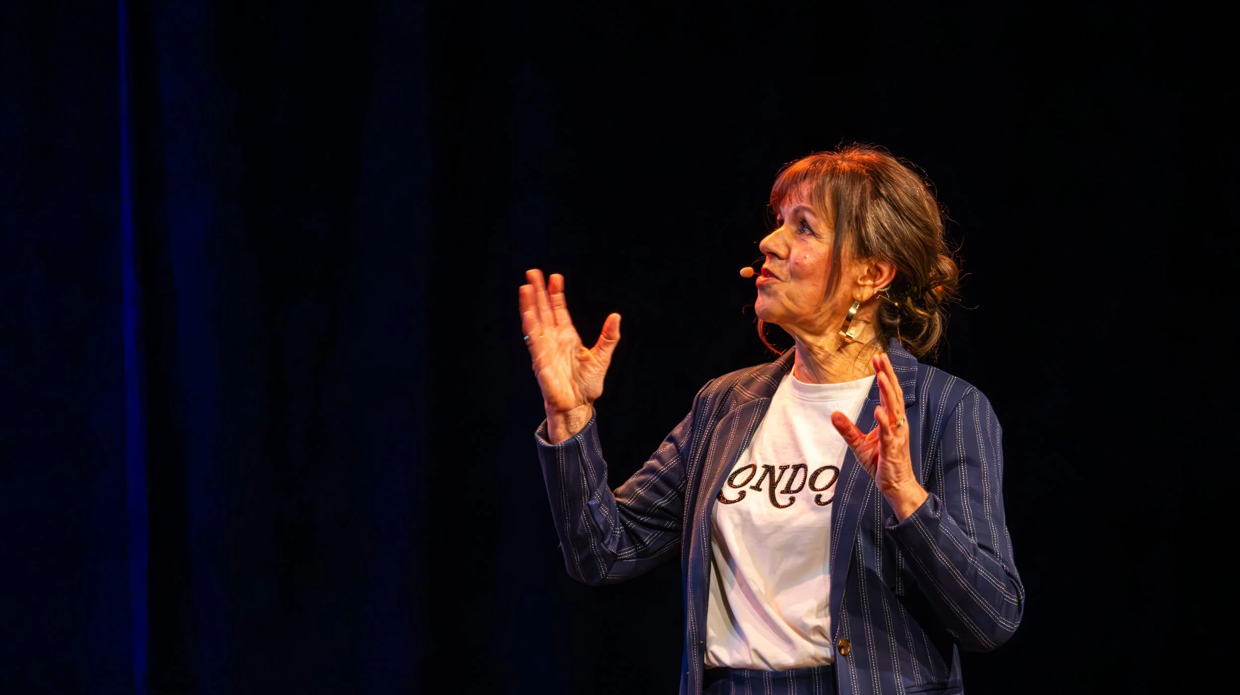 Een oudere vrouw met korte bruin haar spreekt tijdens een presentatie of optreden op een podium, gekleed in een blauwe streepjasje en een witte T-shirt.