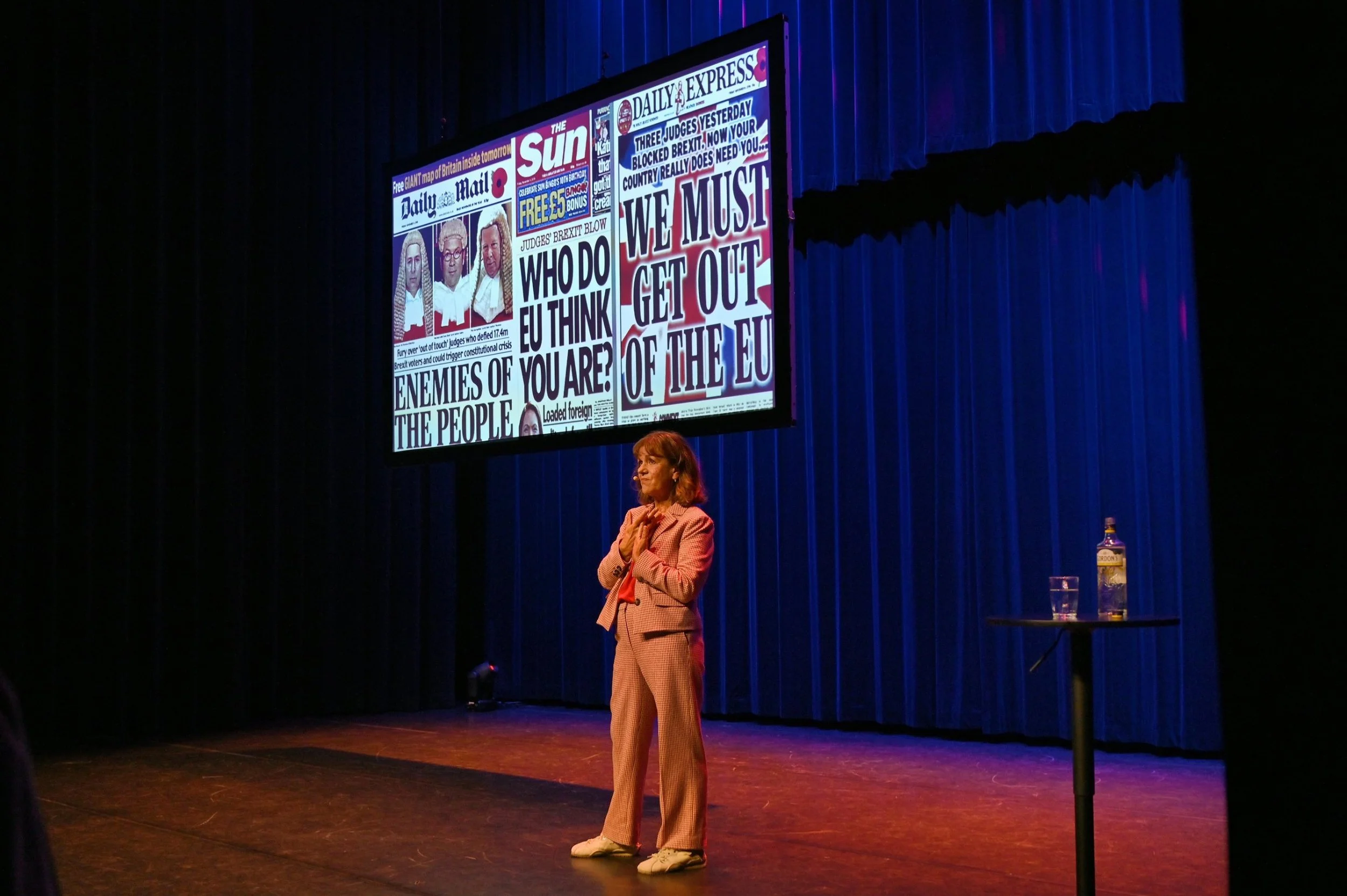 Lia van Bekhoven op het podium tijdens Leven (en lijden) met de Britten, met Britse krantenkoppen over Brexit op het scherm achter haar