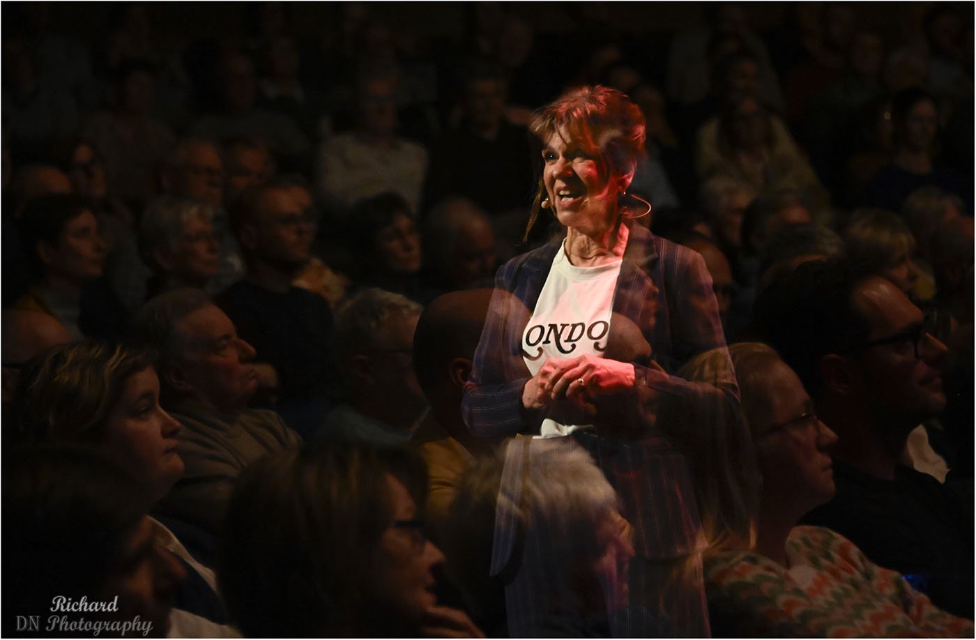 Vrouw op een podium die lacht en publiek dat naar haar kijkt, bij een evenement of optreden.