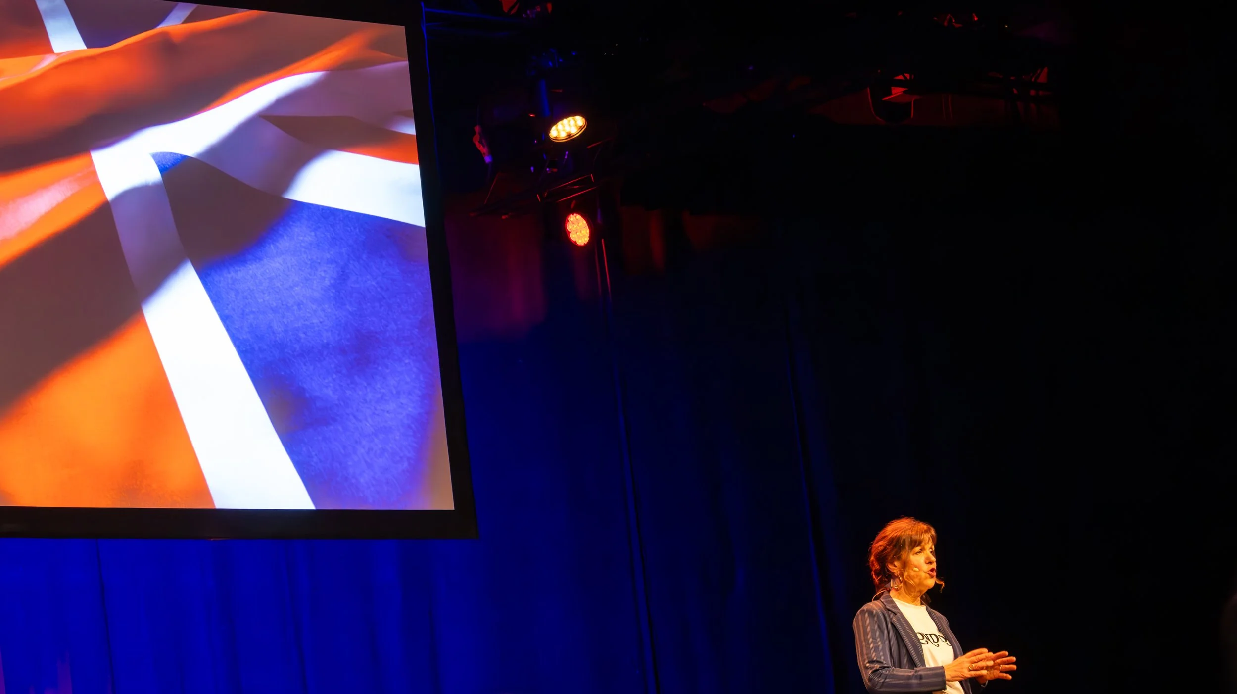 Een vrouw staat op een podium met een grote scherm achter haar dat de Britse vlag toont; er hangen rode lichten en het is donker in de zaal.
