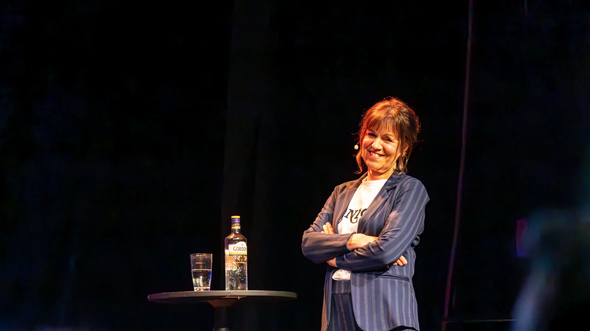 Vrouw met handen gekruist voor haar borst, glimlachend op een podium, met een fles Gordon's gin en een glas op een ronde tafel.