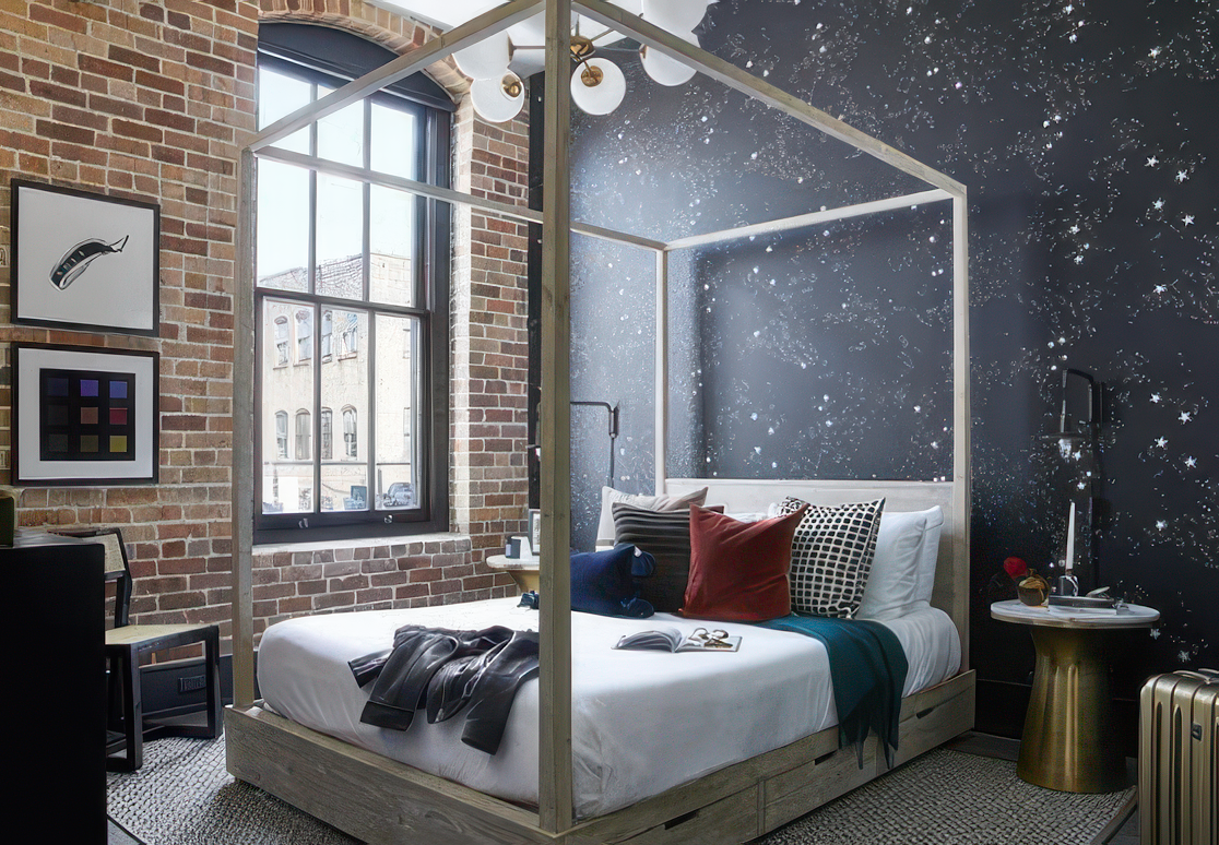 A modern bedroom with a brick wall, large window, and constellation-themed dark blue accent wall. A white bed with a canopy frame is decorated with various pillows, and a black leather jacket is draped over the bed. There are framed artworks on the b
