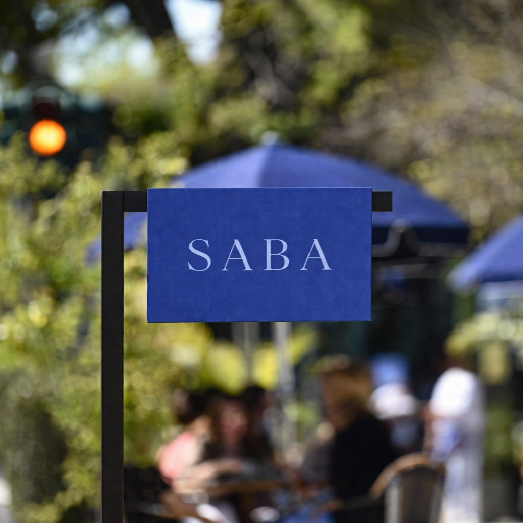 Blue sign with the word 'SABA' placed outdoors with tables and people dining under umbrellas, trees in the background, and a traffic light showing an orange signal.