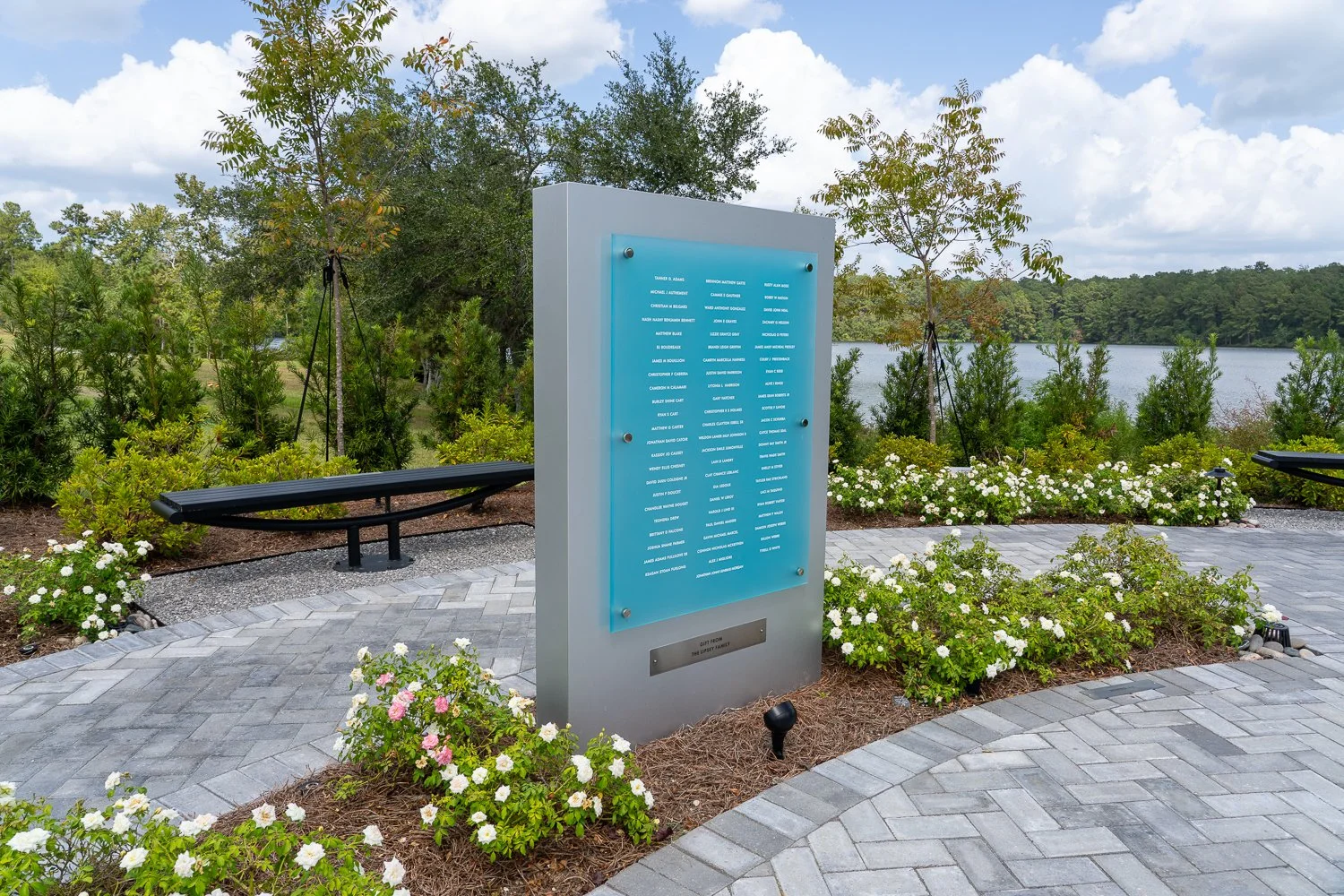 A memorial park with a gray and blue informational plaque, surrounded by white and pink flowers, green bushes, two black benches, and trees, with a lake and cloudy sky in the background.