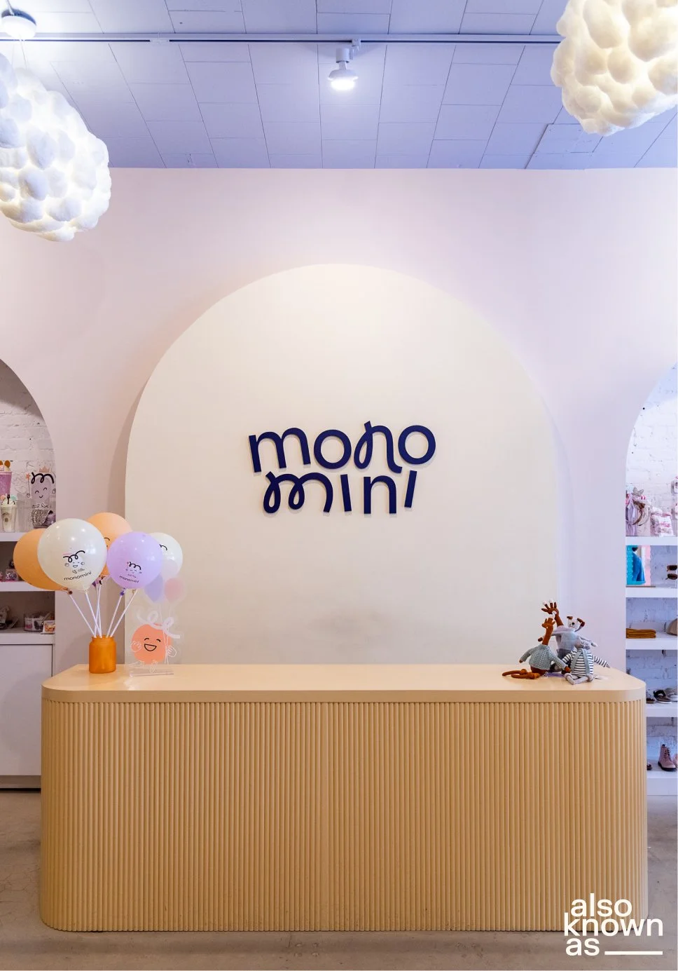 Interior of a retail store with a beige ribbed counter, a white wall with a large cream-colored circle, and the sign 'mono mini' in dark blue letters. There are pastel balloons on the counter, a small orange vase, and plush toys of reindeer and a rab