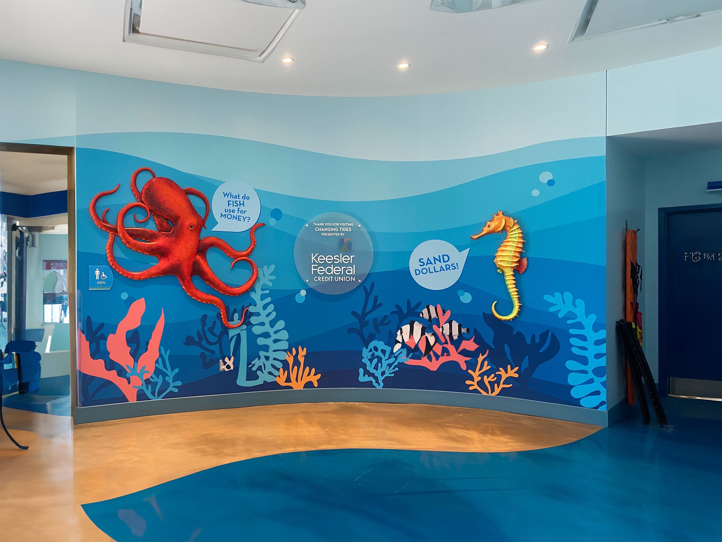 Colorful underwater-themed mural with an octopus and seahorse, featuring signs asking 'What do fish use for money?' and 'Sand dollars!', at Keesler Federal Credit Union.