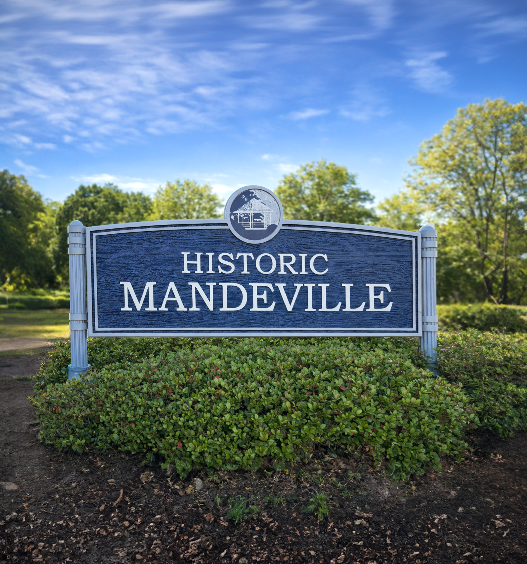 A sign reading "Historic Mandeville" with a gazebo icon at the top, surrounded by green bushes and trees under a partly cloudy blue sky.