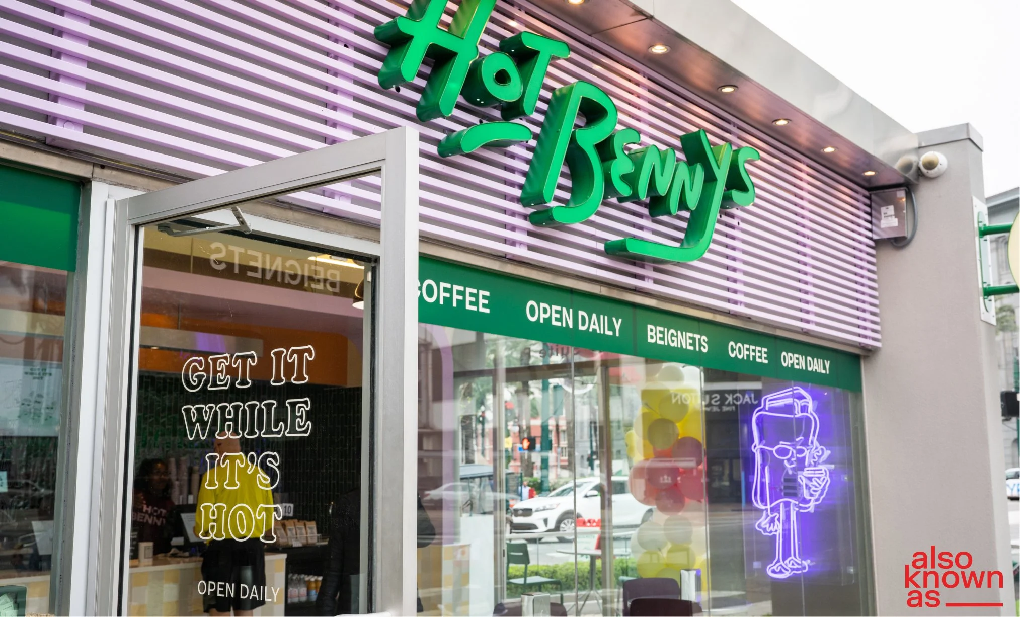 Exterior view of a Benny's Hot Dogs restaurant with neon signs, a glass door, and promotional text on the window. The restaurant offers coffee, beignets, and is open daily. Neon lights include a character holding a hot dog and a drink.