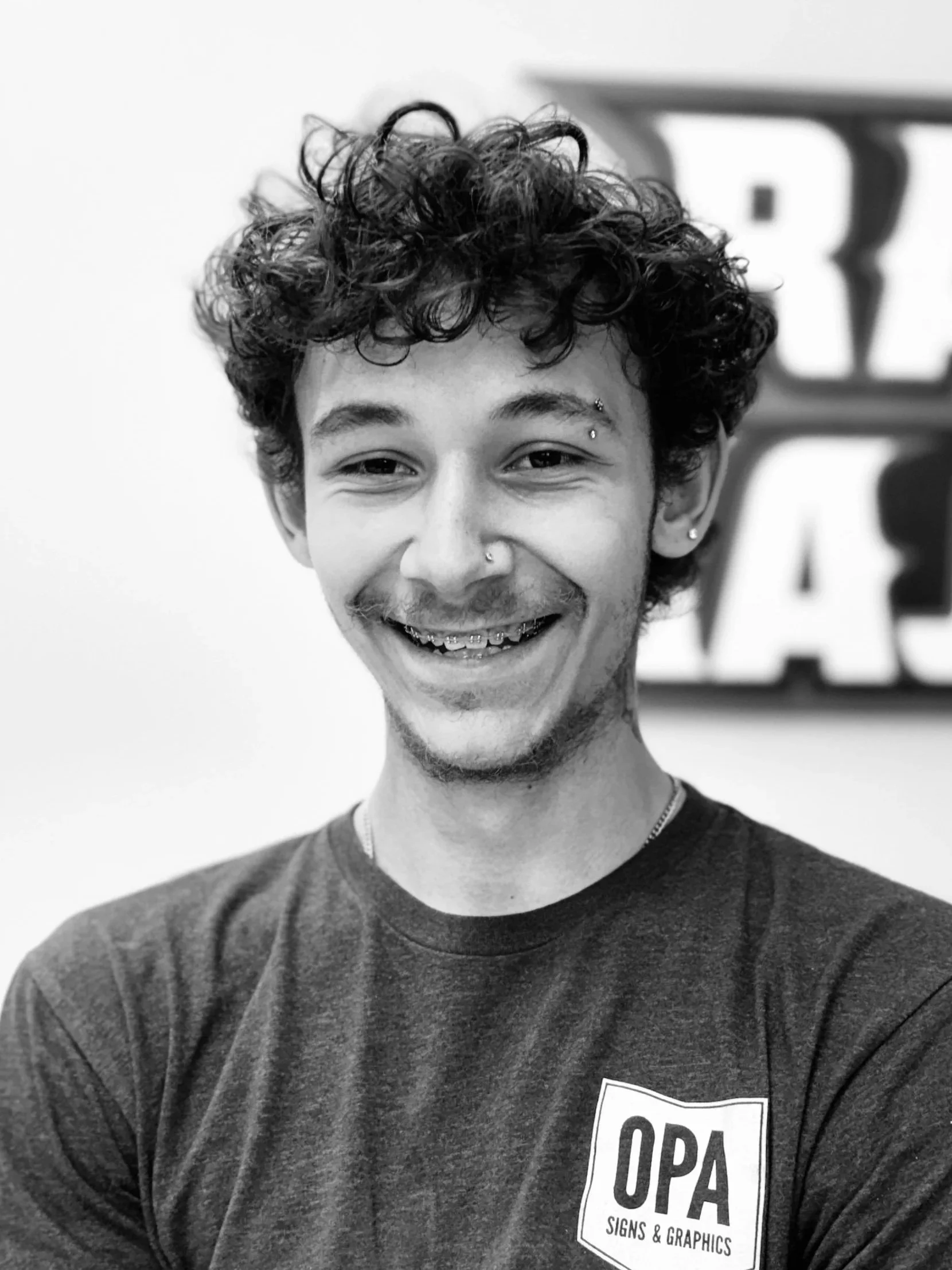A young man with curly dark hair, multiple facial piercings, and a broad smile, wearing a dark T-shirt with a logo that reads "OPA Signs & Graphics," standing in front of a sign in the background.