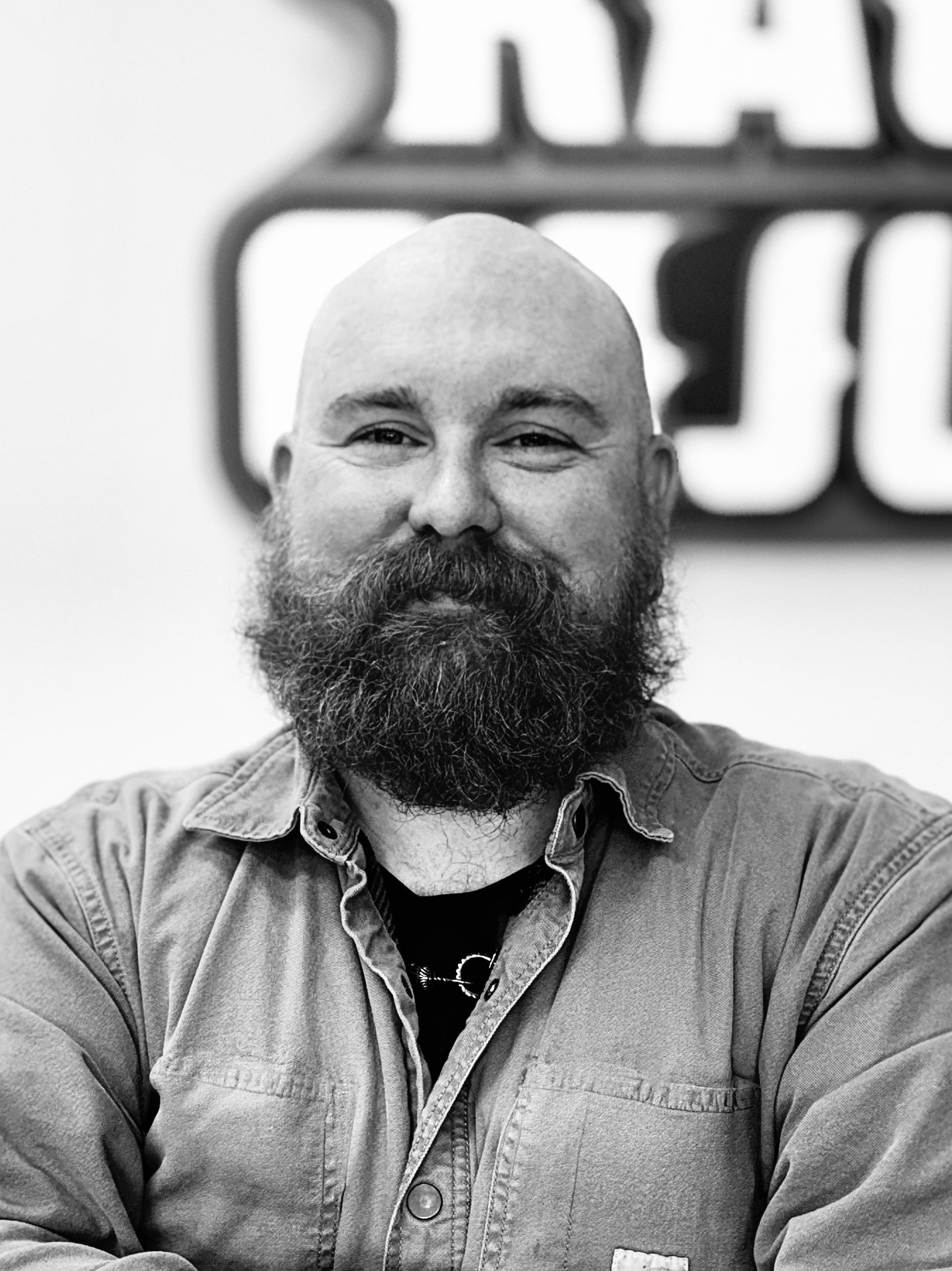 A man with a beard and a mustache, smiling, with a bald head, wearing a button-up shirt, sitting in front of a blurred background with large letters.
