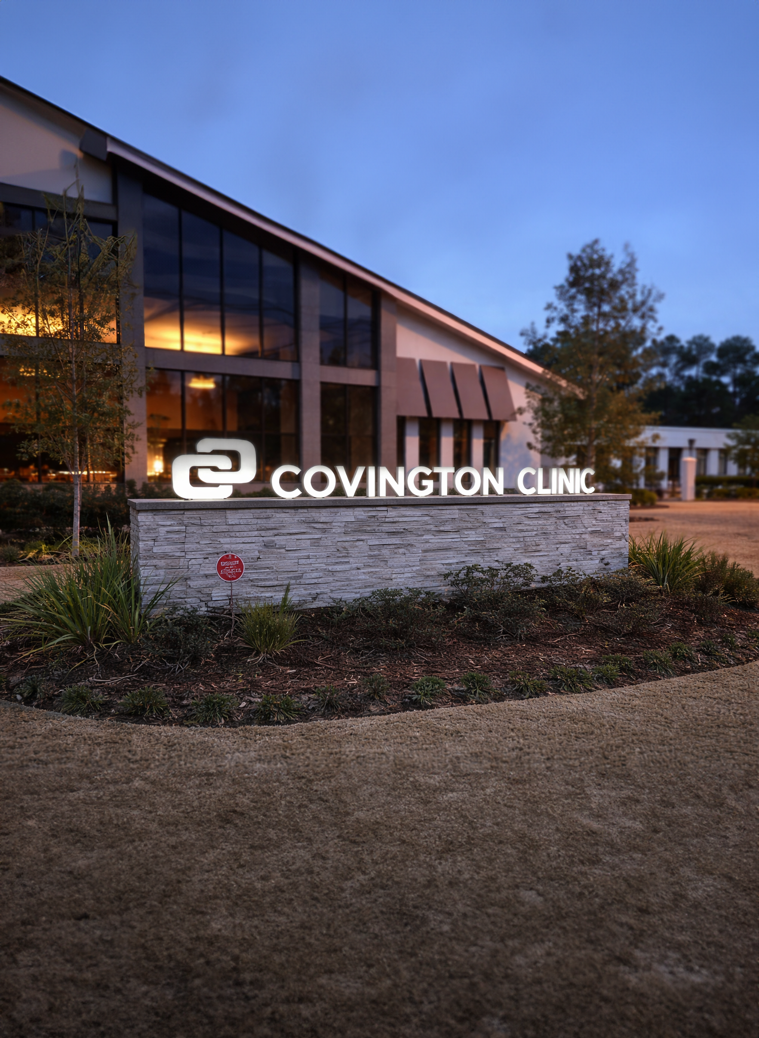 Exterior of Covington Clinic building at dusk, with large glass windows illuminated from inside, modern architecture, landscaped front yard with plants and trees, and a sign displaying the clinic's name.