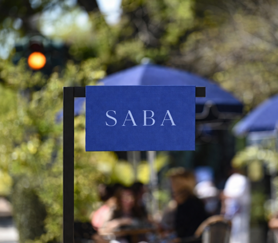 Blue sign with the word on it that says "SABA" in an outdoor setting with people sitting and eating at tables under umbrellas in the background
