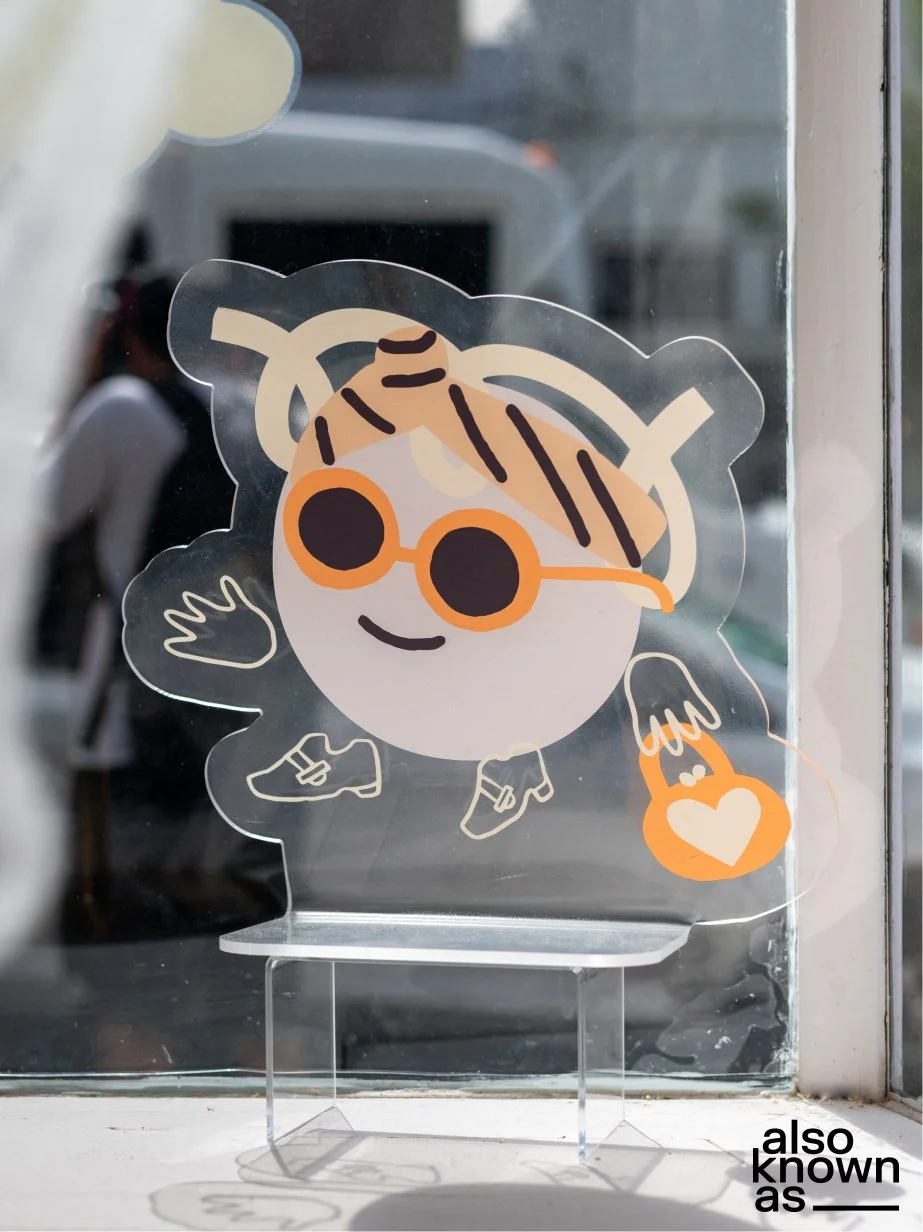 Sticker of a cartoon character with a round face, wearing orange sunglasses, a striped headband, striped shoes, and holding a small handbag with a heart on it, placed on a window with reflections in the background.