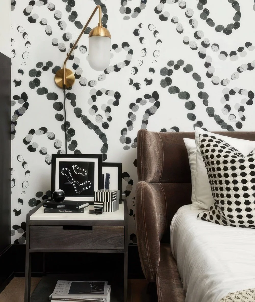 A bedroom with a black and white abstract wallpaper, a brass and glass wall lamp, a brown velvet bed with polka dot pillows, and a white bedside table with black and white framed art, books, and decorative objects.