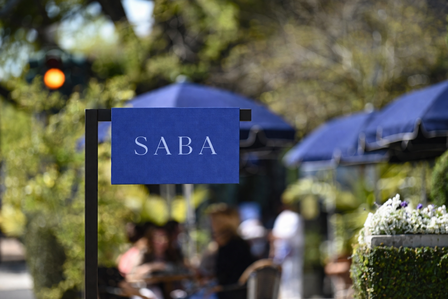 Outdoor restaurant with a blue sign that reads 'SABA', blue umbrellas, and blurred people dining outdoors, with greenery and trees in the background.
