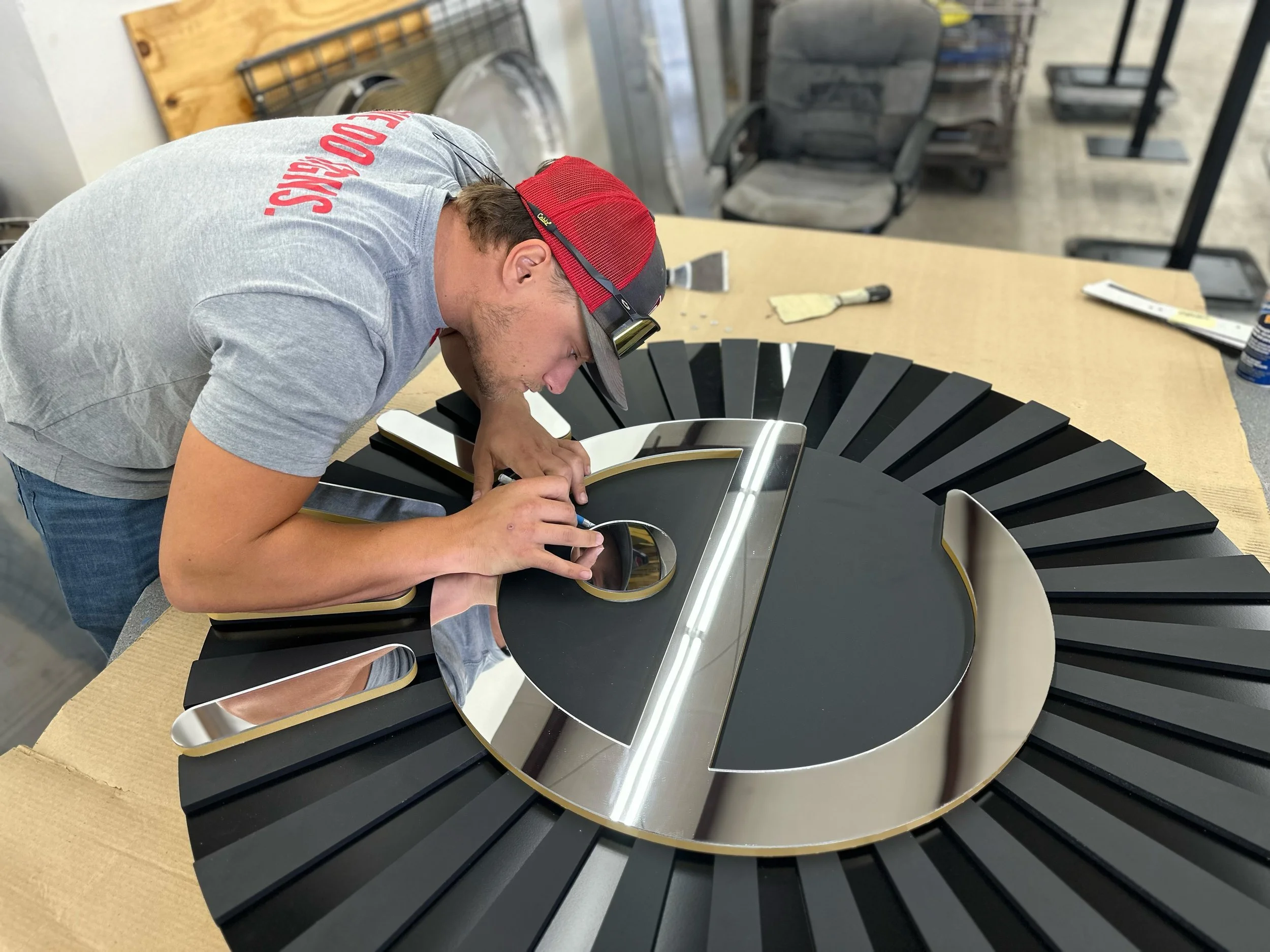 A man in a gray shirt and red cap working on assembling a circular sign with black and metallic gold accents at a worktable.