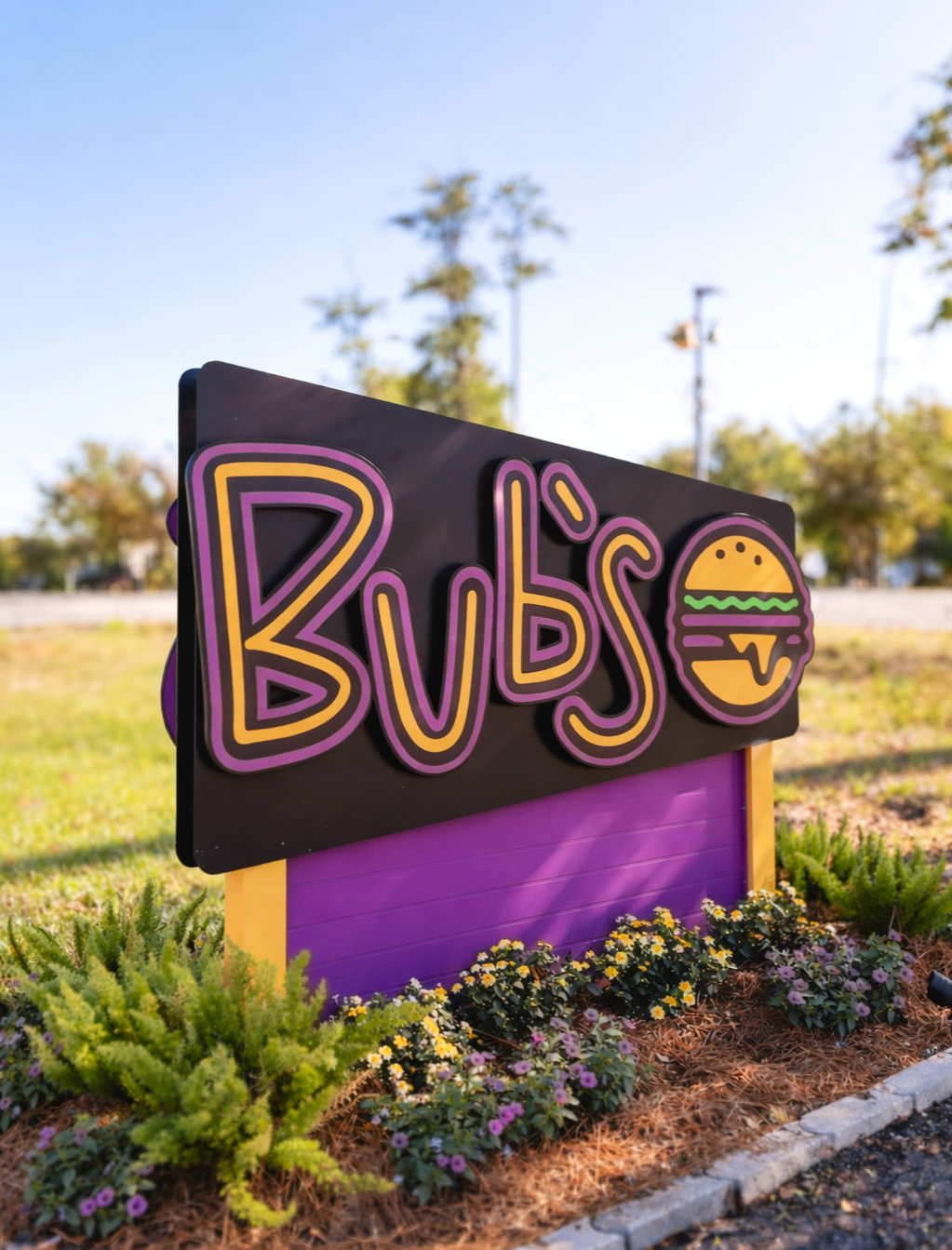 Colorful sign reading 'BUB'S' with a burger icon, located outdoors among small flowers and plants.
