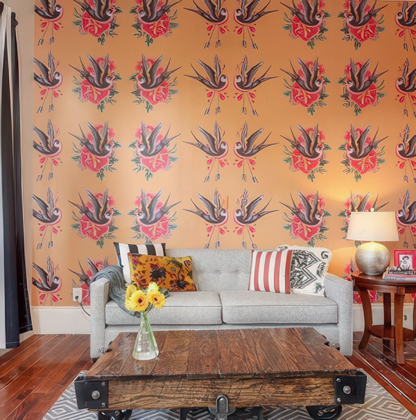 Living room with wallpaper of roosters and flowers, gray couch with floral and striped pillows, wooden coffee table, vase of yellow flowers, and lamp on side table.