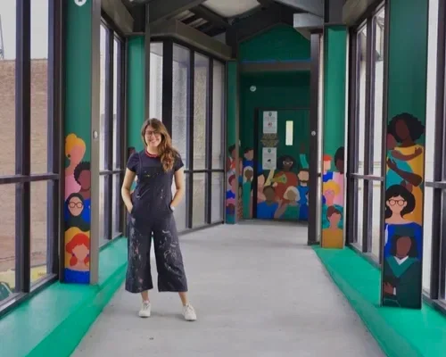 A woman standing inside a glass-enclosed walkway with colorful murals of diverse people on the walls.
