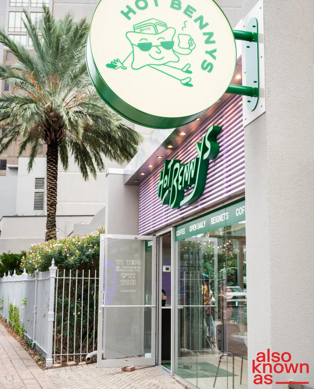 Outdoor view of a Benny's restaurant with a green circular sign featuring a cartoon bread character wearing sunglasses, holding a coffee mug, and the restaurant's name. The building has a purple and green sign, glass entrance doors, and a palm tree i