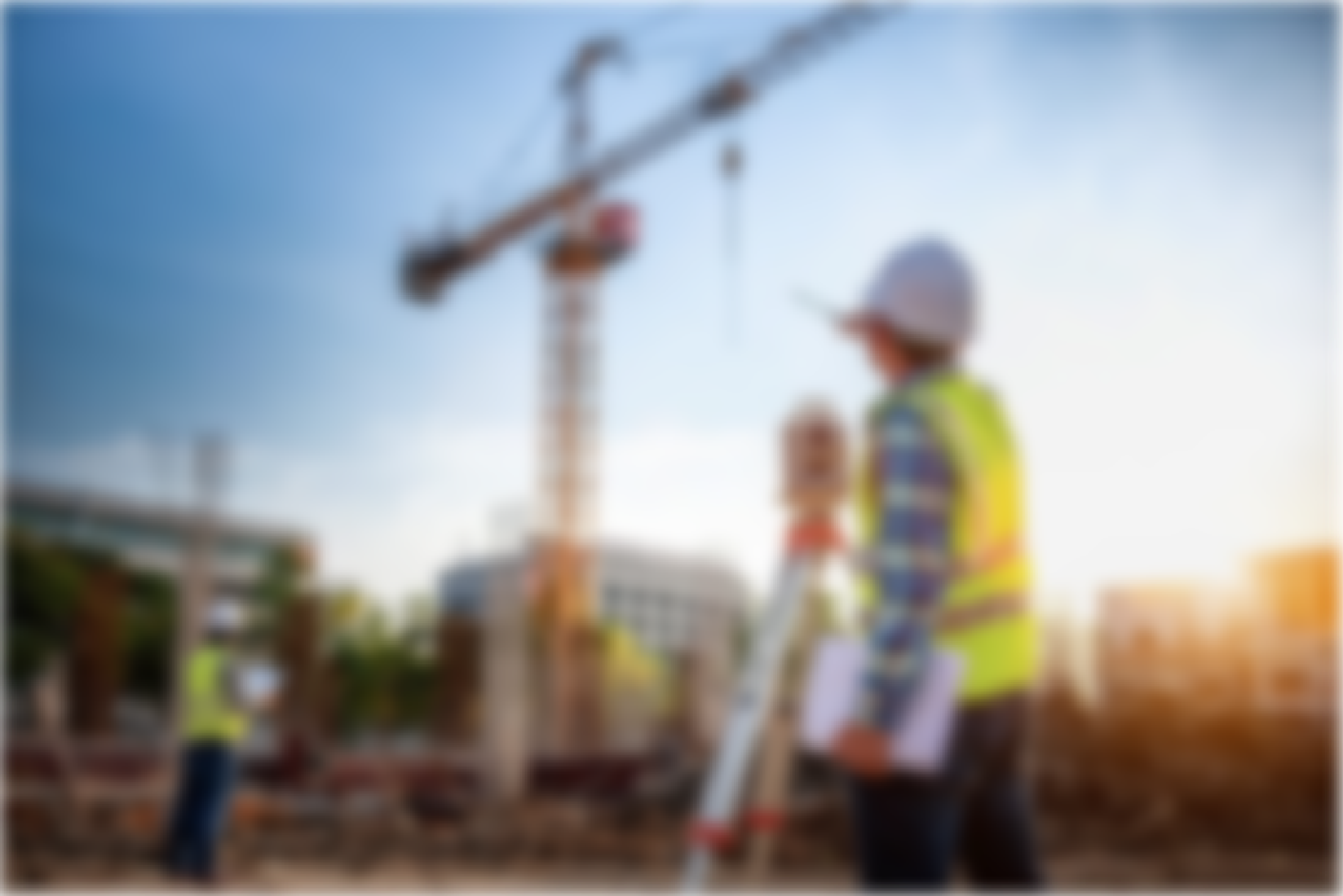A construction worker with a white helmet and yellow safety vest holding a tablet, standing near a construction site with the crane in the background during sunset.