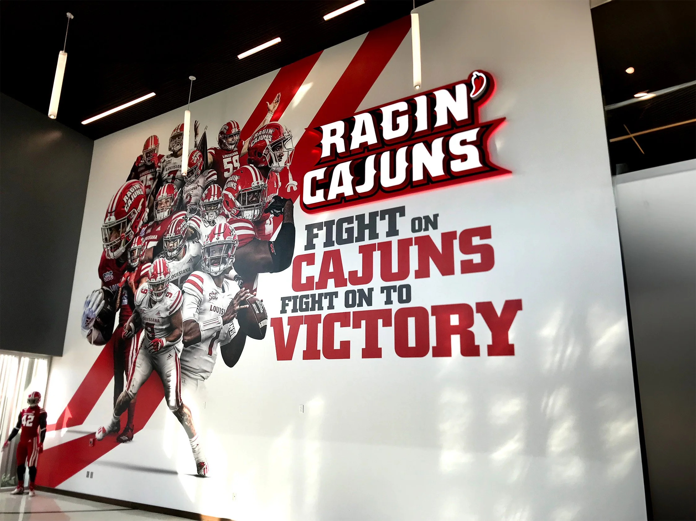 A large wall mural featuring football players in red and white uniforms and the text 'Ragin Cajuns, Fight on Cajuns, Fight on to Victory' with team logo.