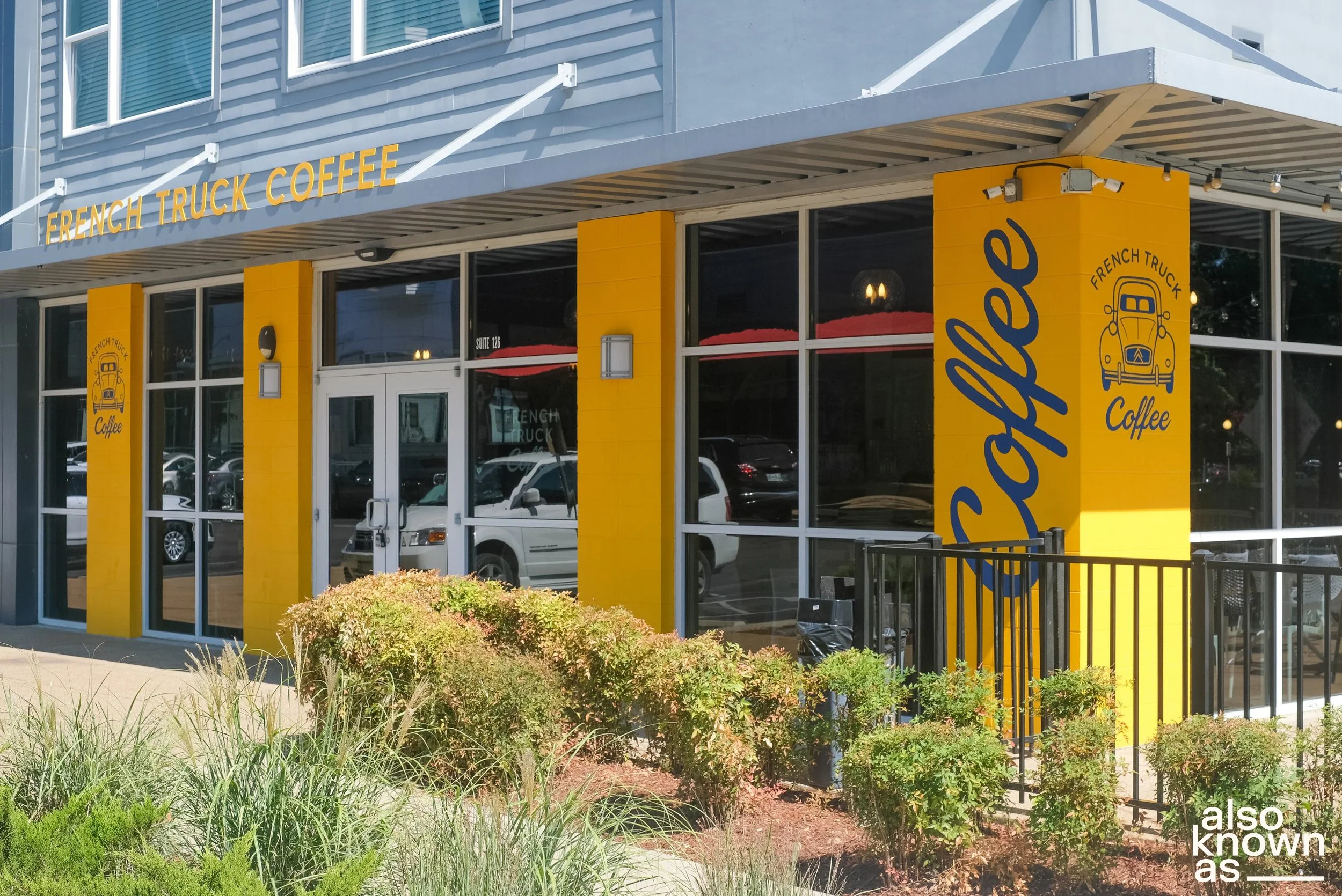 Exterior of a coffee shop named French Truck Coffee with yellow pillars and large glass windows, outdoor seating area, and surrounding bushes.