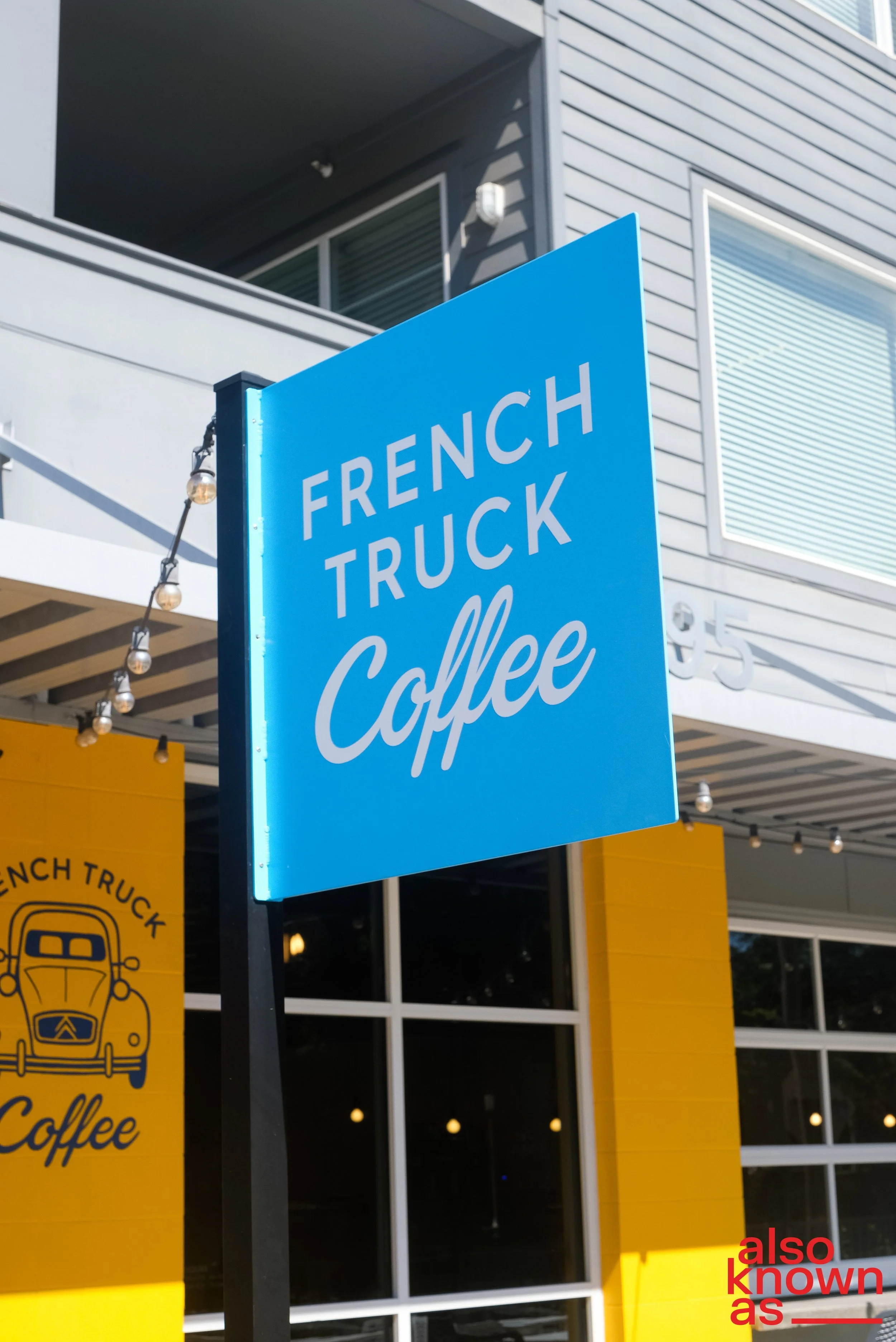 Blue sign for French Truck Coffee outside a modern building with yellow accent walls, large windows, and string lights.