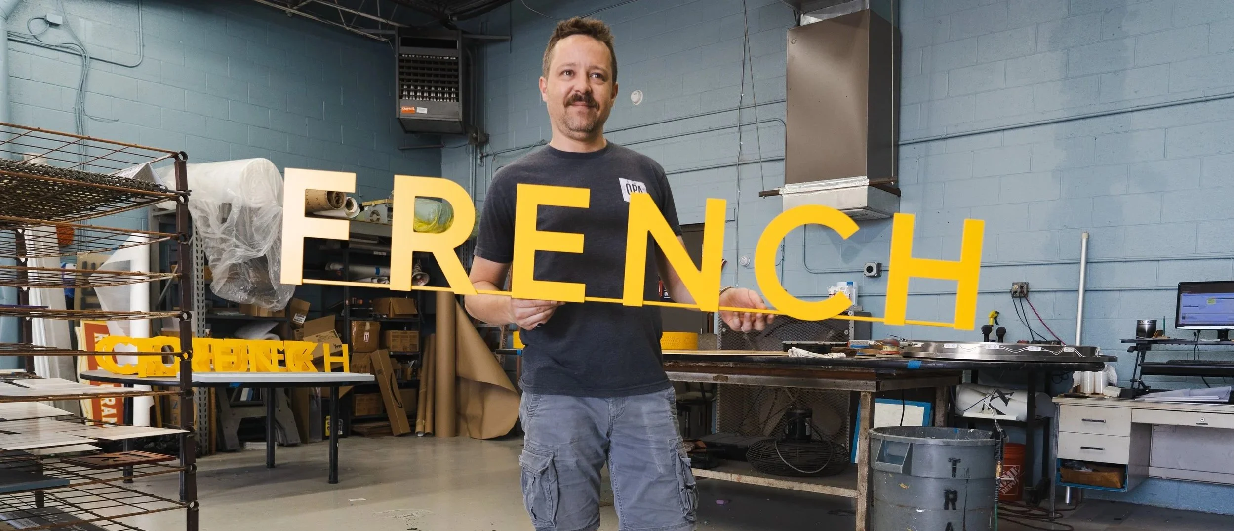 A man standing in a workshop holding a yellow sign that spells "FRENCH" in capital letters.