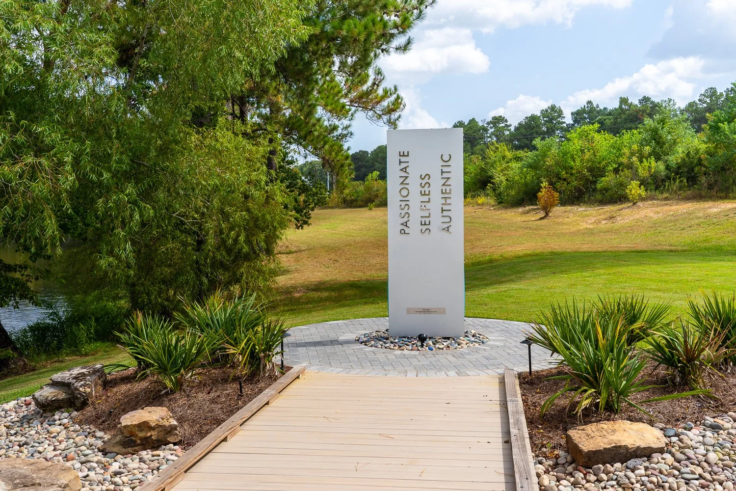 A white vertical sign with the words "Passionate, Selfless, Authentic" standing on a circular brick platform in a grassy outdoor area surrounded by trees and shrubs, with a cloudy sky above.