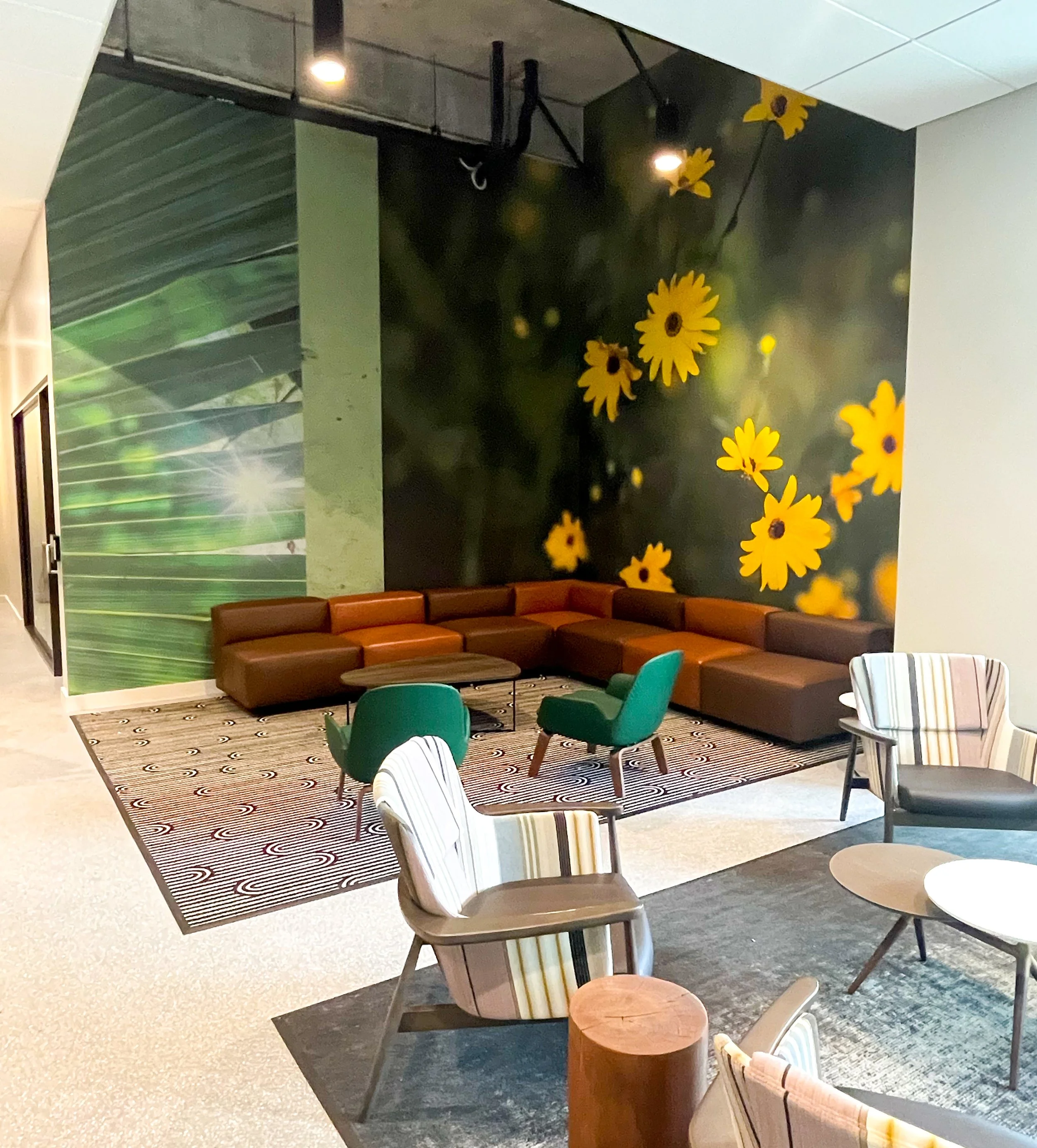 Lobby area with a large wall mural of yellow flowers and green foliage, brown sectional sofa, two green chairs, striped armchair, and small round tables on a patterned rug.