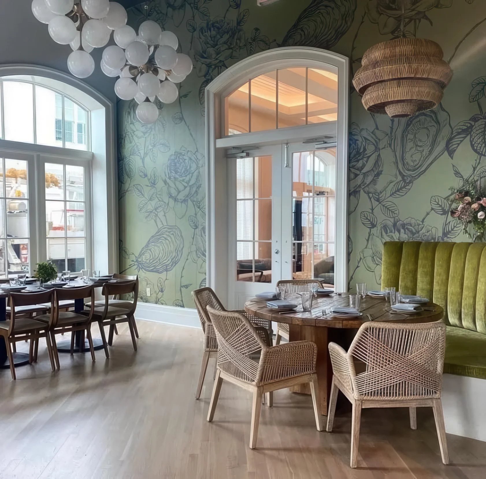 Interior of a restaurant with a large window, a white door with glass panes, and decorative floral wall art. There is a hanging chandelier with white globes, a green velvet booth, and a round wooden table set with plates, glasses, and utensils. Wicke